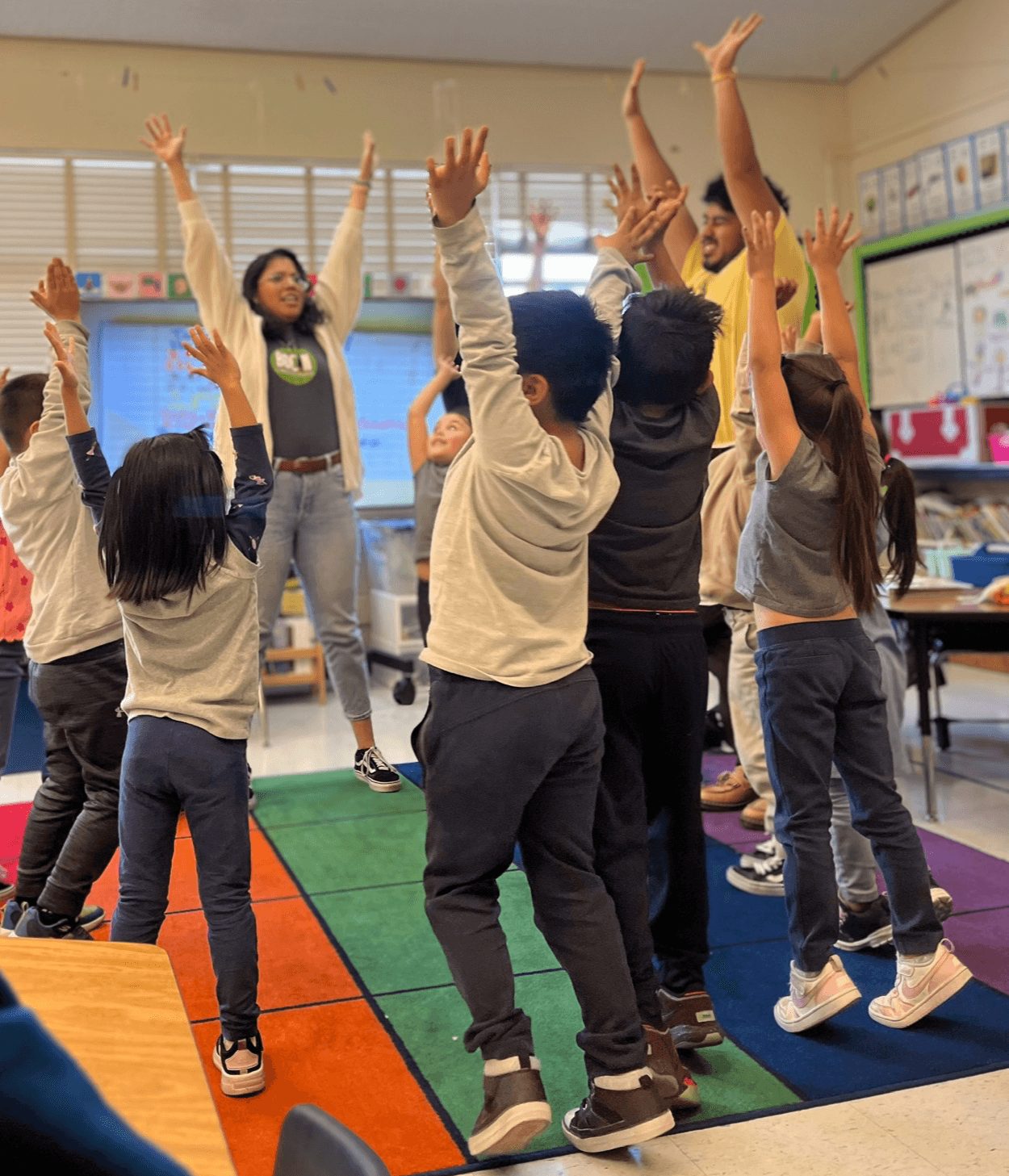 Children in a classroom raising their hands, with a teacher leading activity, colorful classroom environment, and educational posters.