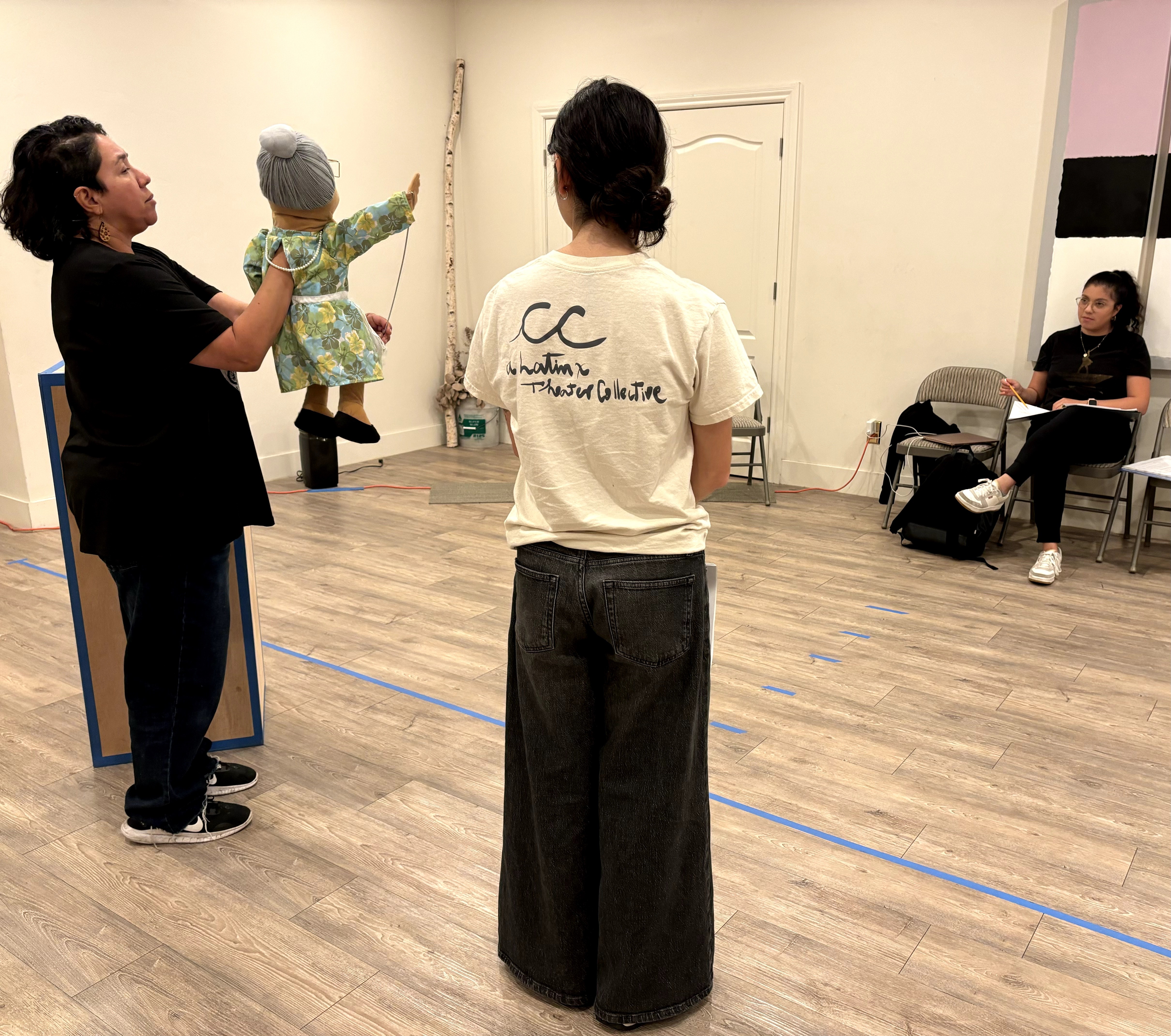 Three women and a child in a room with wooden floor, one woman holding the child, another facing away, a woman sitting on a chair.