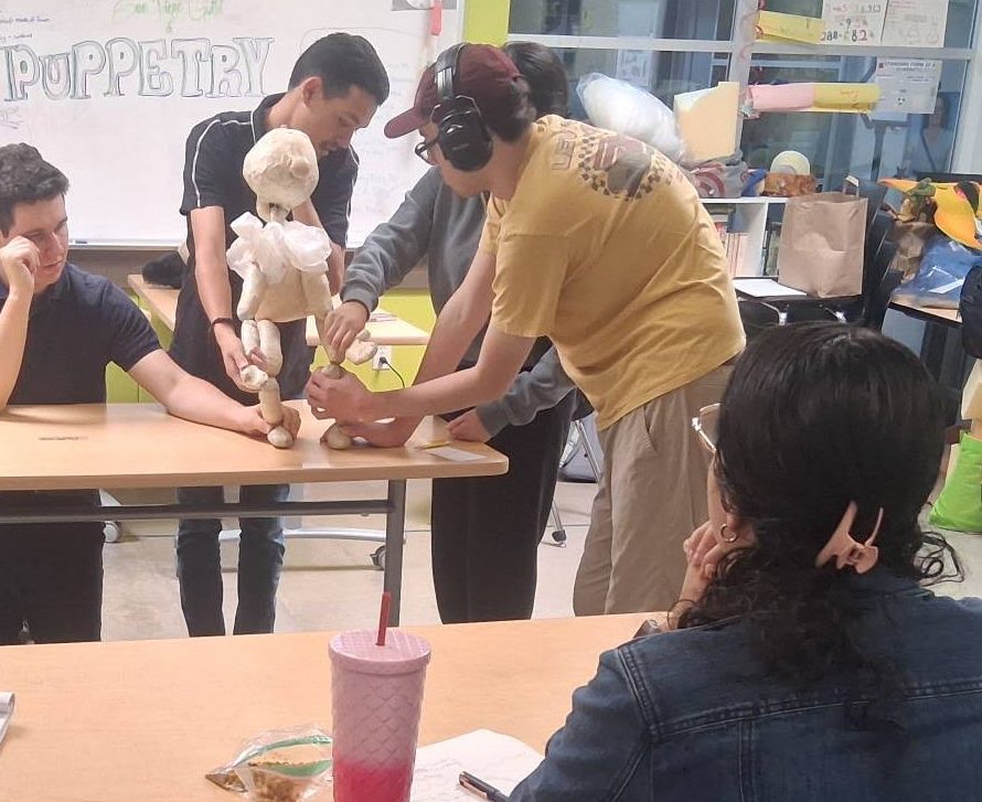 Group of people around a table, one person wearing headphones, engaging with a teddy bear, in a classroom setting.