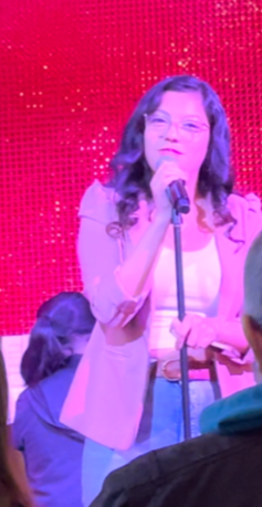 Young woman with glasses and wavy hair speaking into a microphone on stage with a red background.