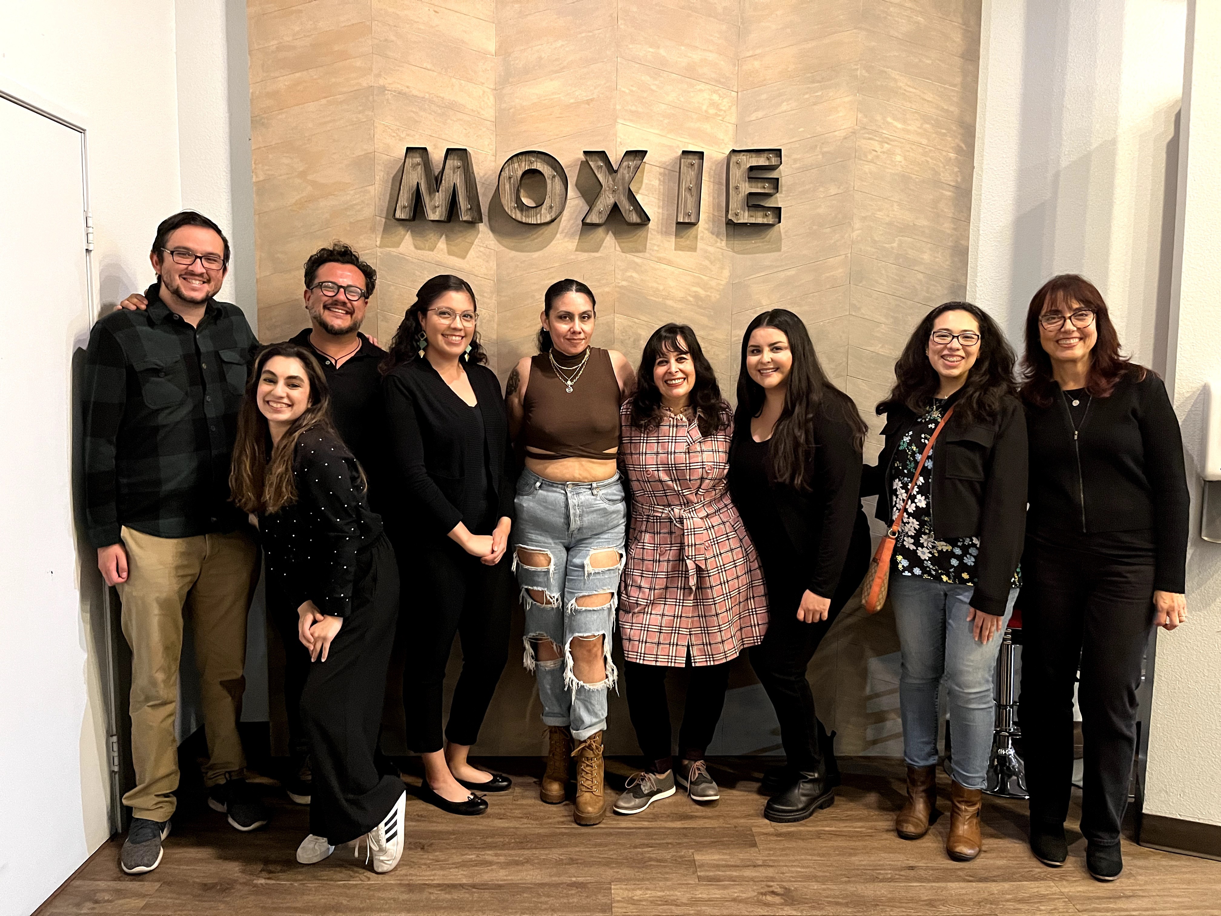 Group of nine people standing indoors in front of a wall with the word 'MOXIE' on it, smiling and posing for the photo.