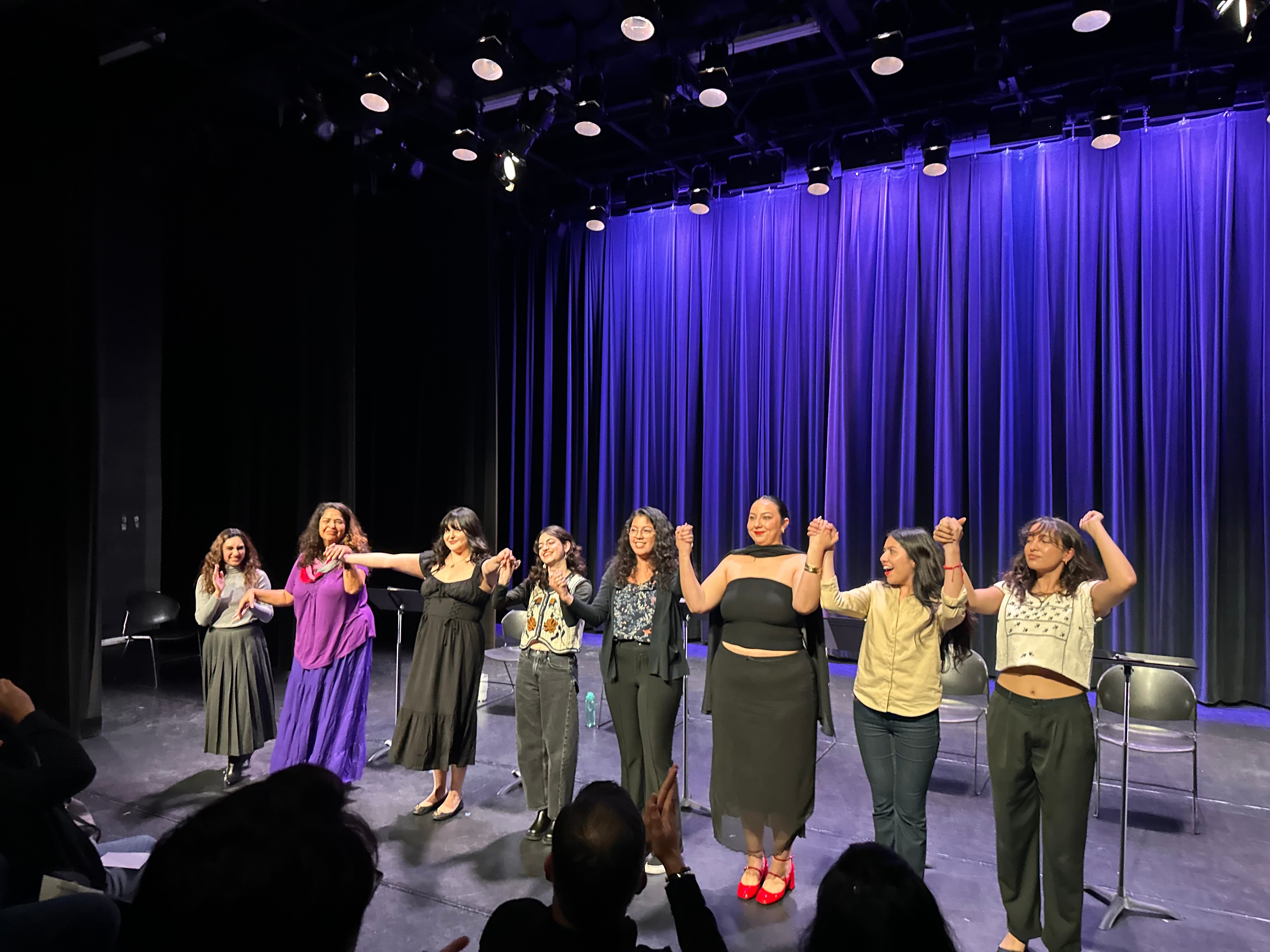 Group of nine women standing on stage with blue curtains, holding hands and raising arms, in a performance or event.