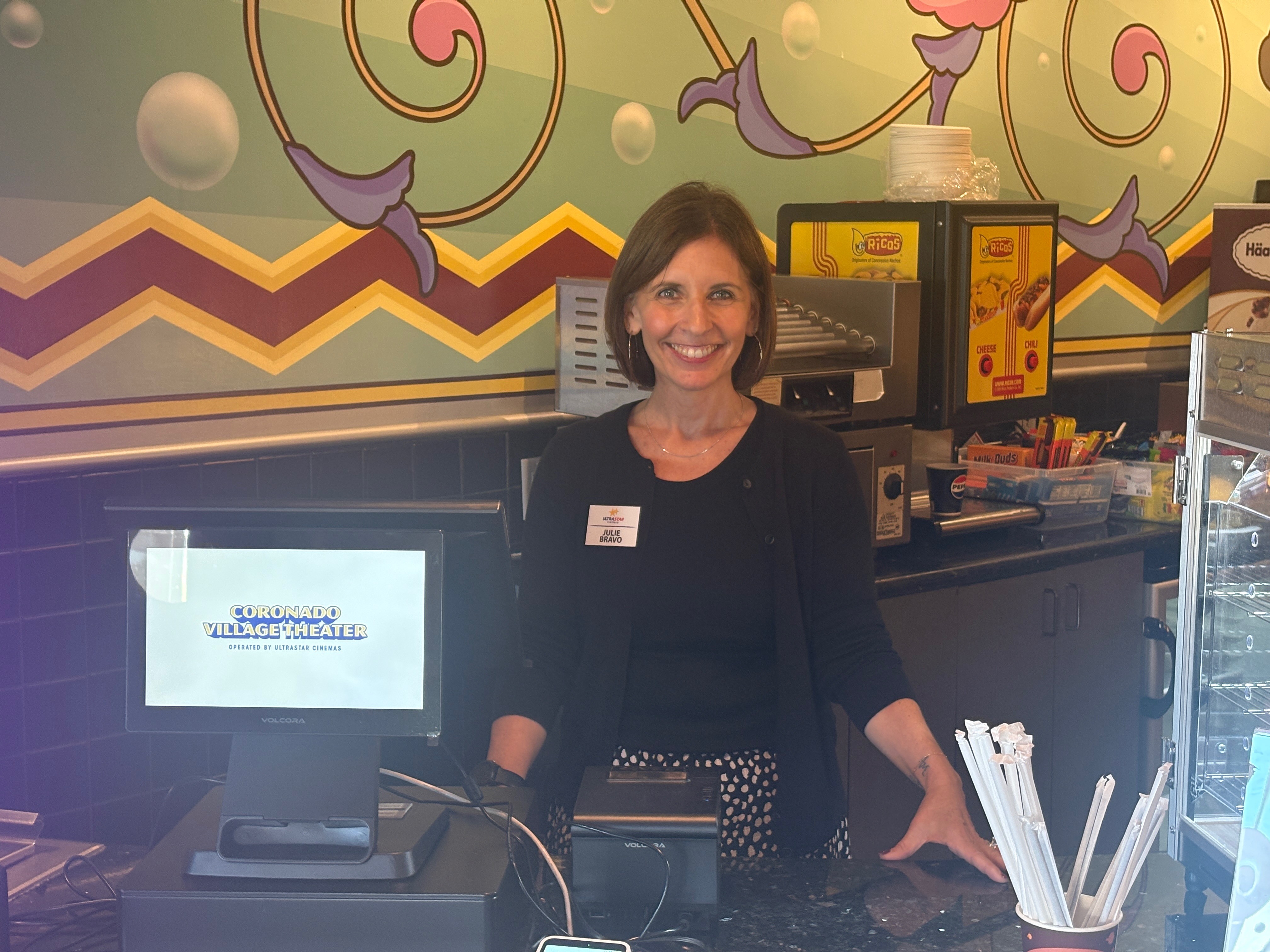 Smiling woman standing behind a counter in a colorful, decorated environment with a computer and kitchen equipment.