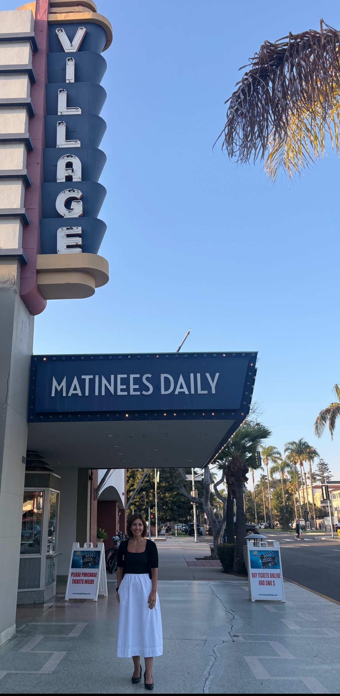 Person standing on sidewalk in front of a building with a sign reading 'VILLAGE' and a marquee that says 'MATINEES DAILY'.