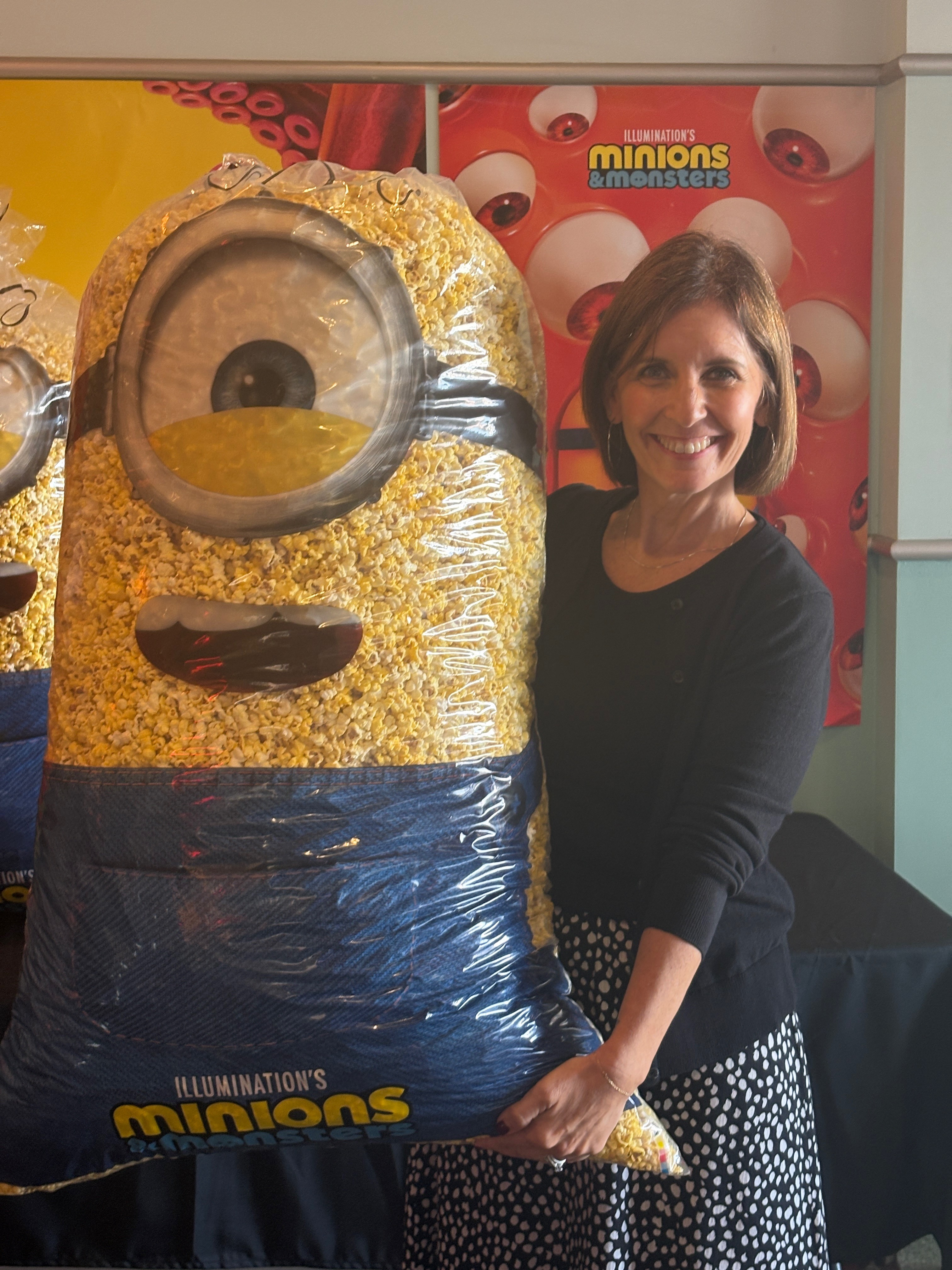 Woman standing next to a large Minions character figure at an event, smiling, with Minions poster in background.