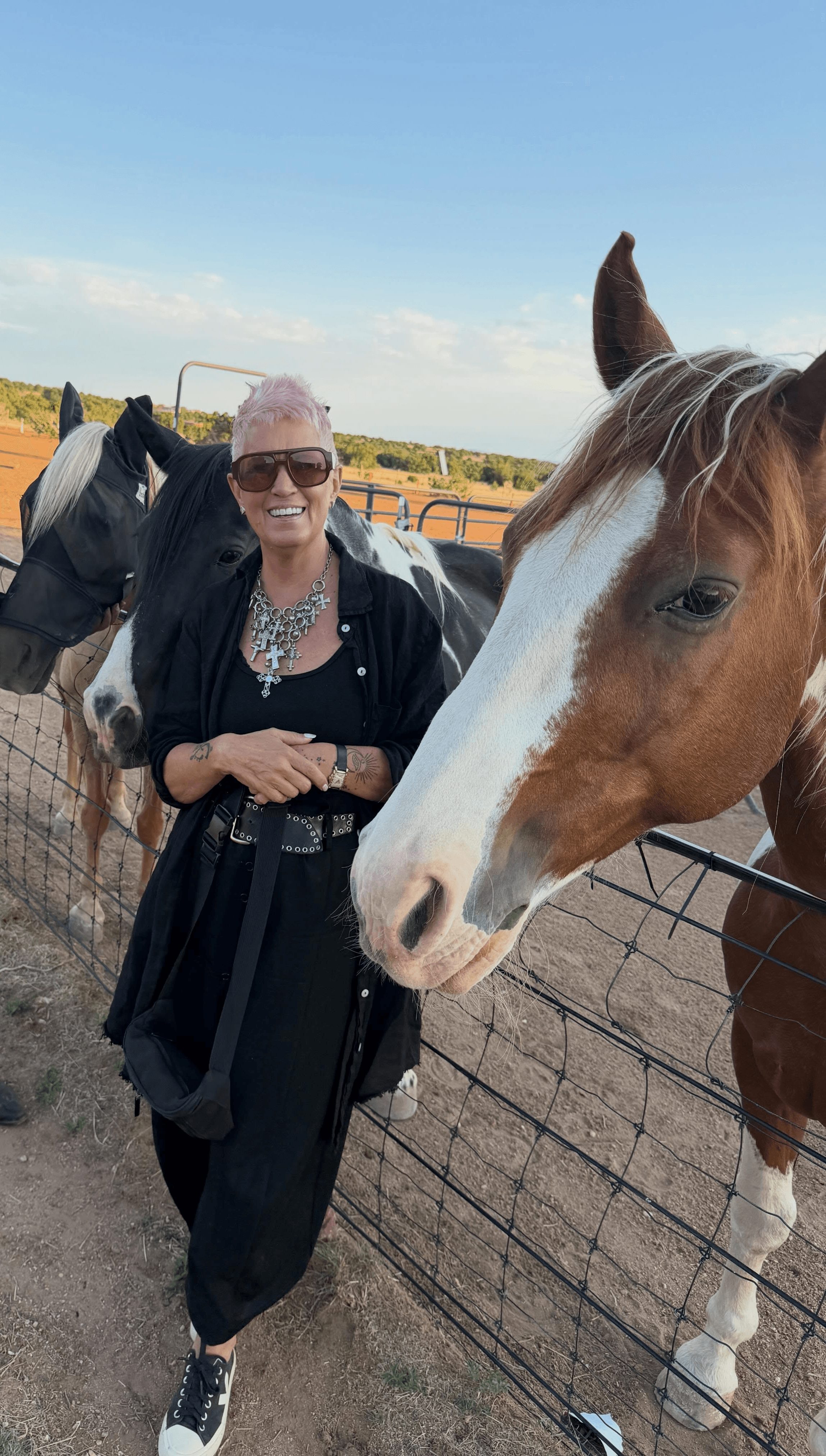 Woman with sunglasses smiling next to a brown and white horse at a fenced outdoor area.