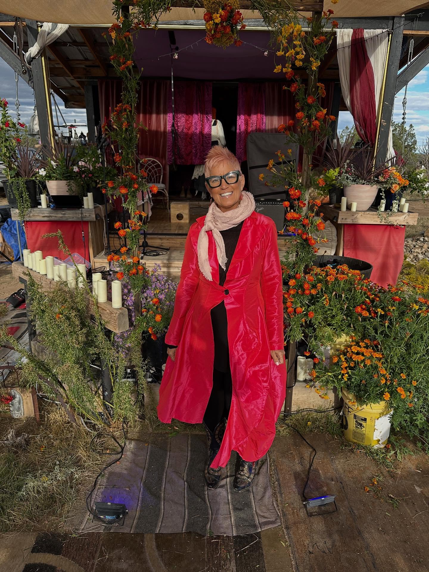 Woman with glasses and scarf standing in front of decorated floral display, wearing a red coat and black outfit.
