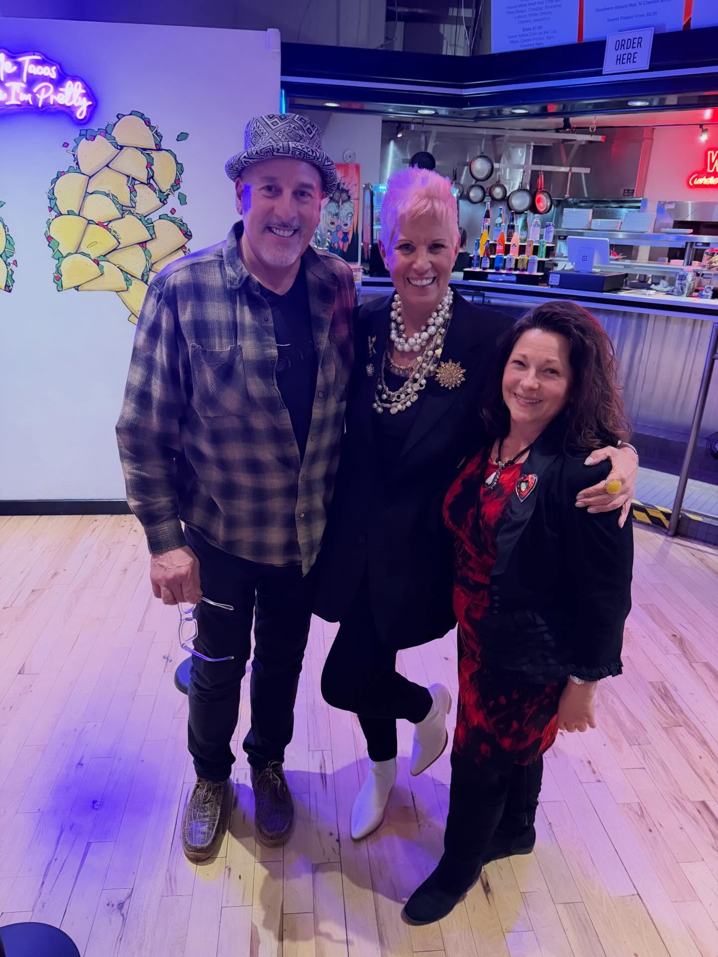 Three people standing together indoors, smiling, with a colorful background and wooden floor.