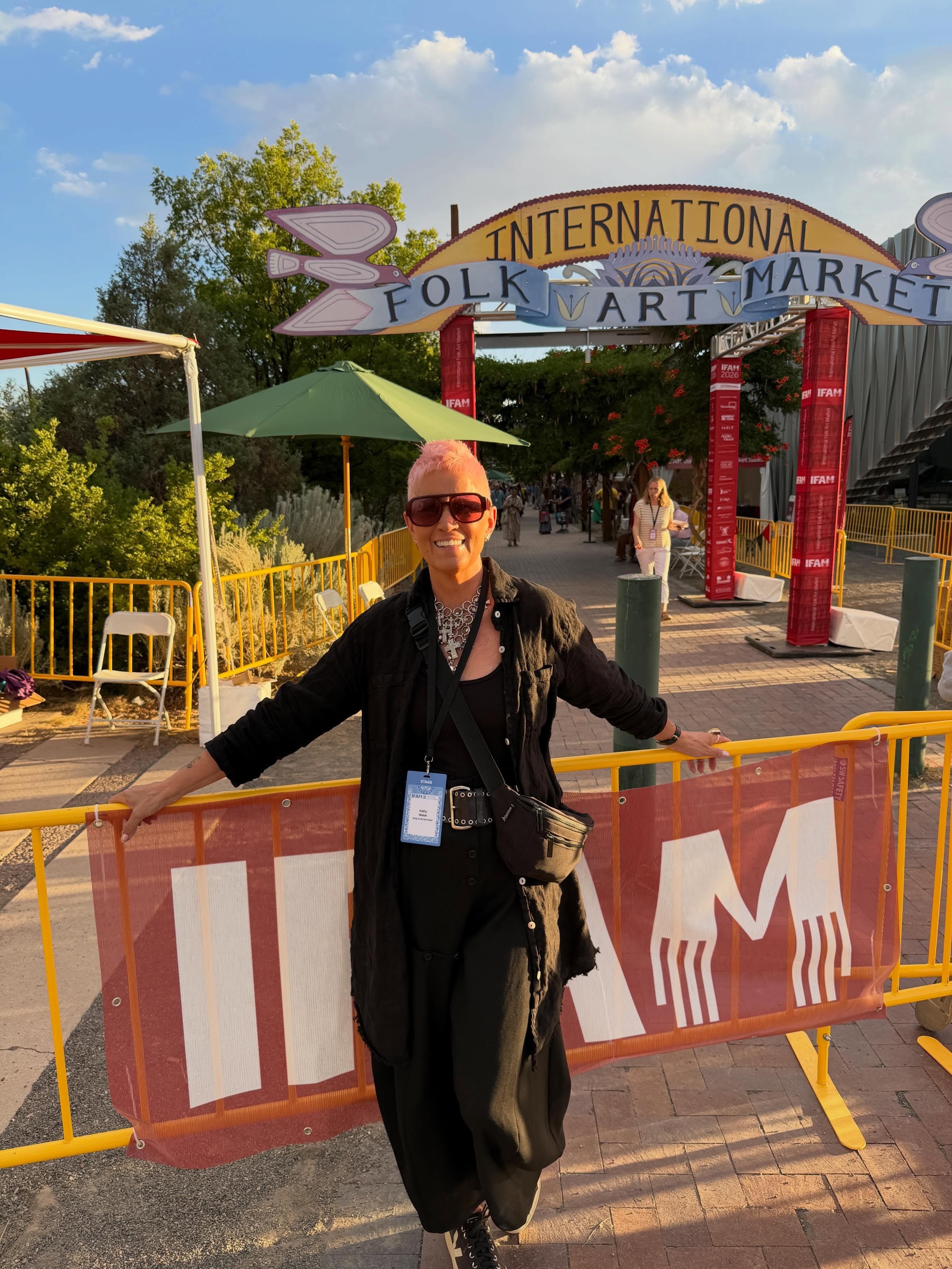 Person standing outdoors at a market entrance with a sign reading 'International Folk Market,' smiling, wearing sunglasses and black clothing.
