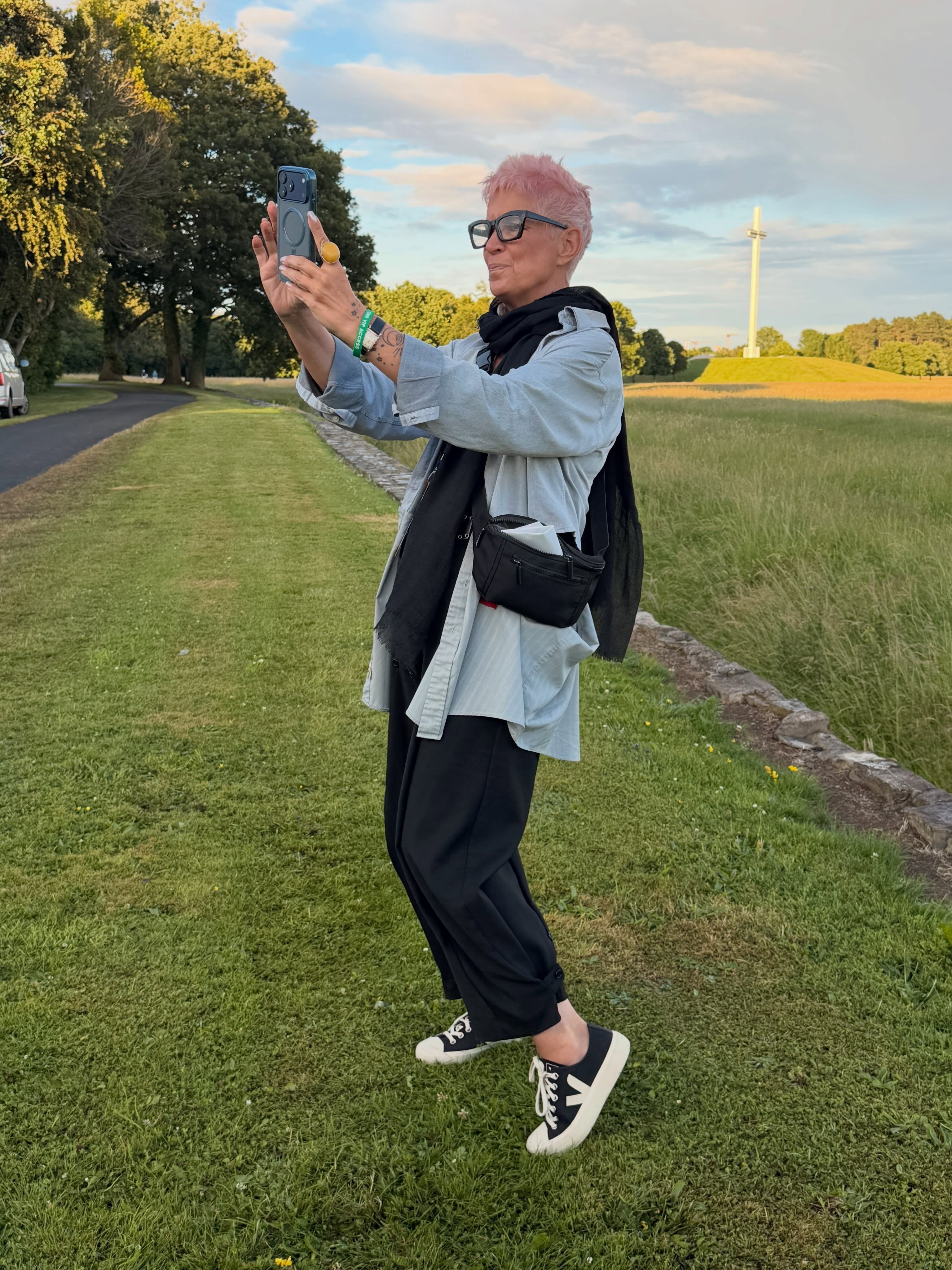 Person with pink hair and glasses taking a selfie outdoors on a grassy path, wearing a light jacket, black pants, and sneakers.