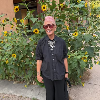 Person with pink hair and sunglasses standing in front of sunflower plants, smiling, wearing a black shirt and pants.