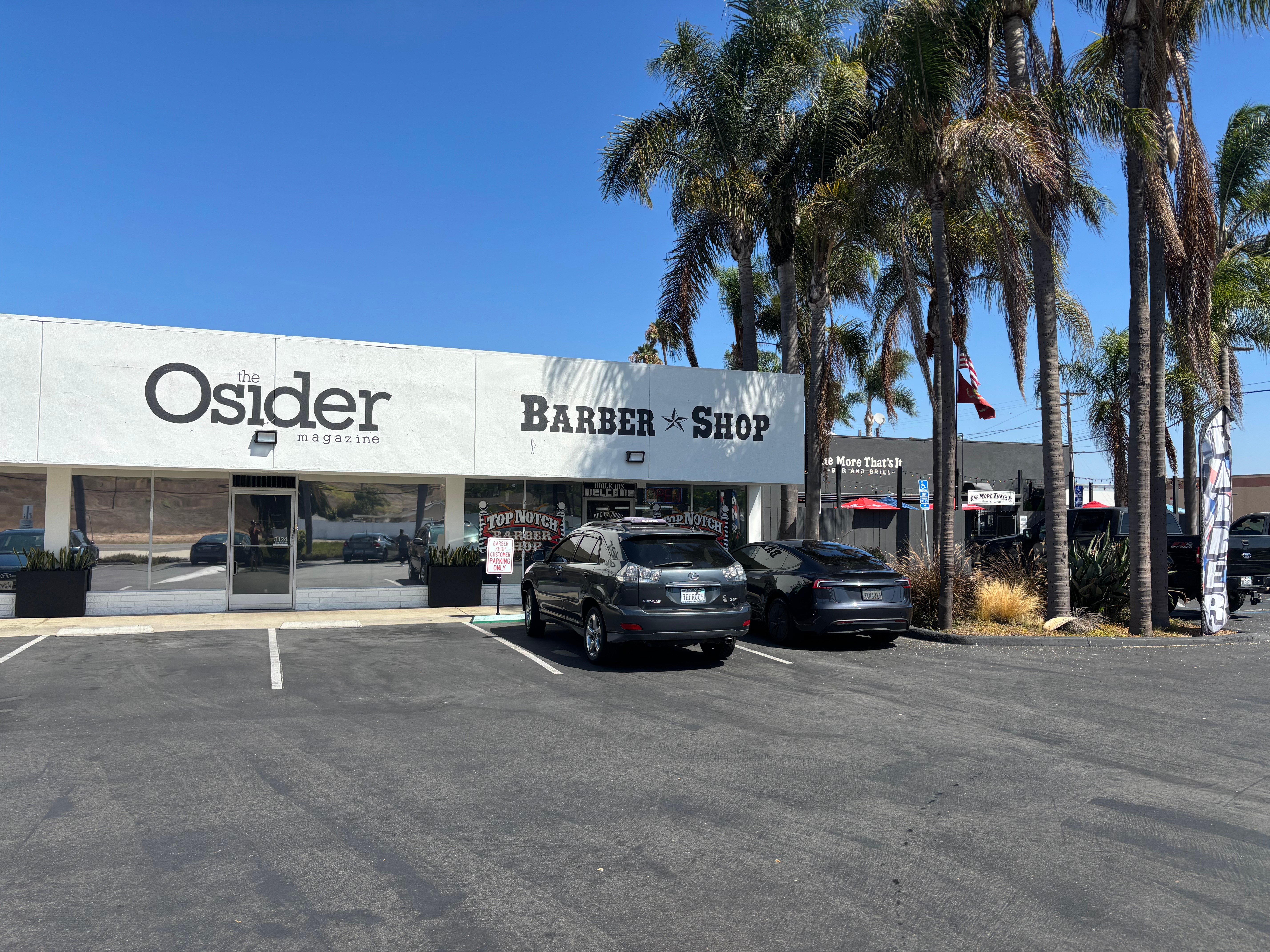 Parking lot in front of a building with signs for Osider and Barber Shop, with palm trees and parked cars.