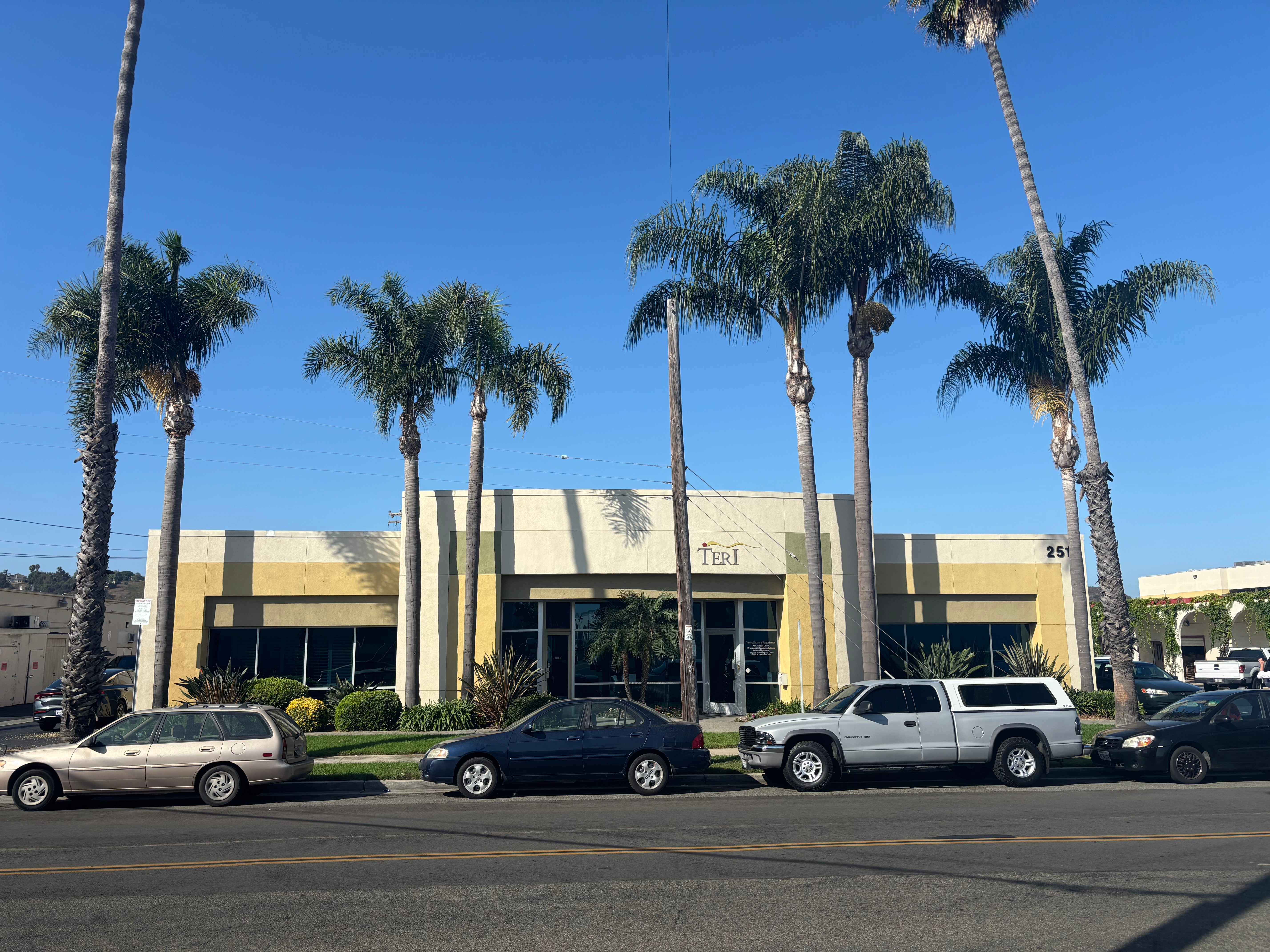 A building with palm trees in front, parked cars along the street, and a clear blue sky.