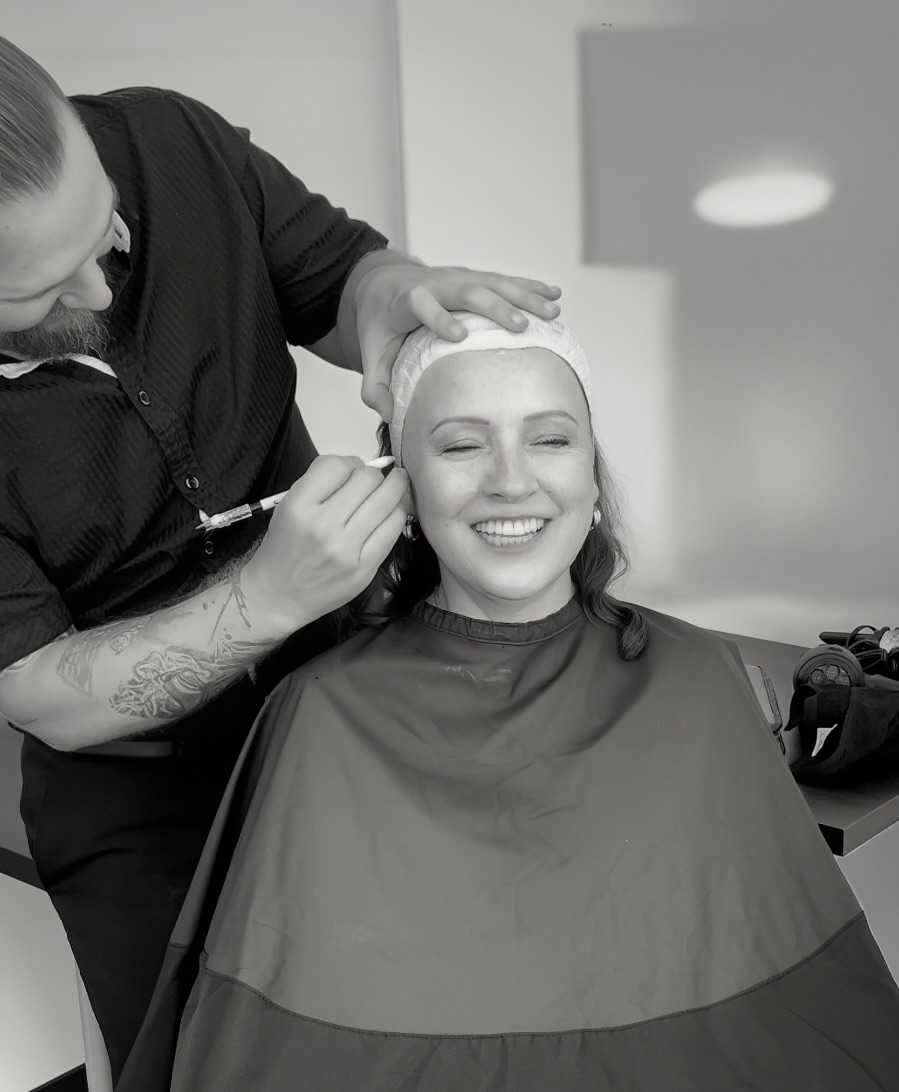 Woman smiling with eyes closed, receiving makeup application from a makeup artist, seated in a salon chair, in black and white.
