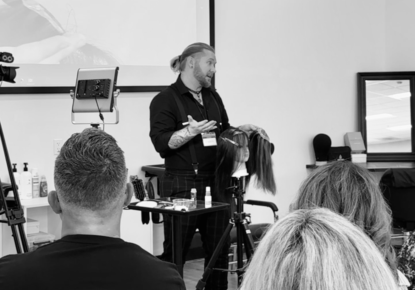 Instructor demonstrating hair styling techniques to a group in a classroom setting, with hairdressing tools on table.