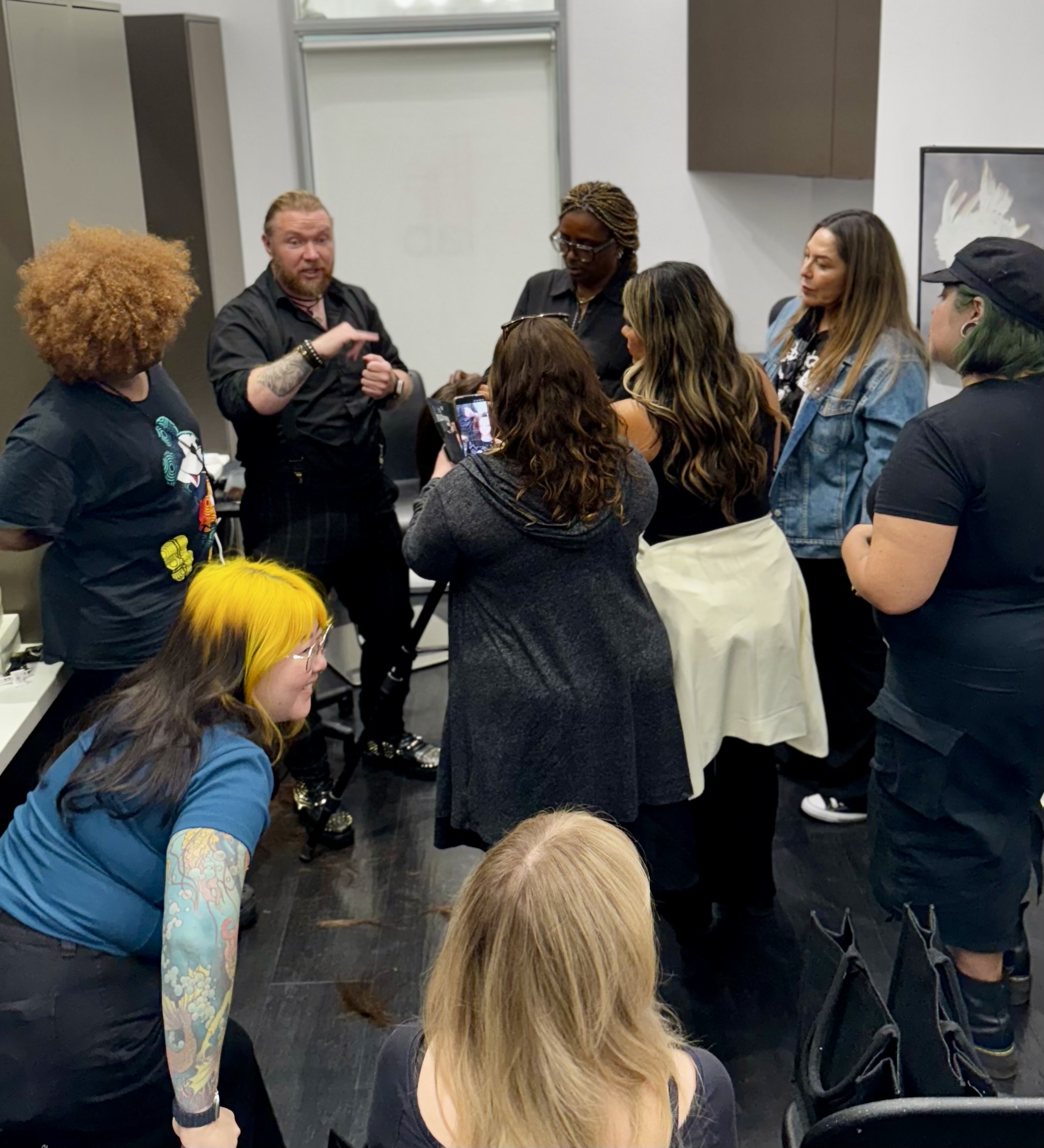 Group of people gathered indoors, listening to a man speaking, with diverse hairstyles and clothing, some taking photos.