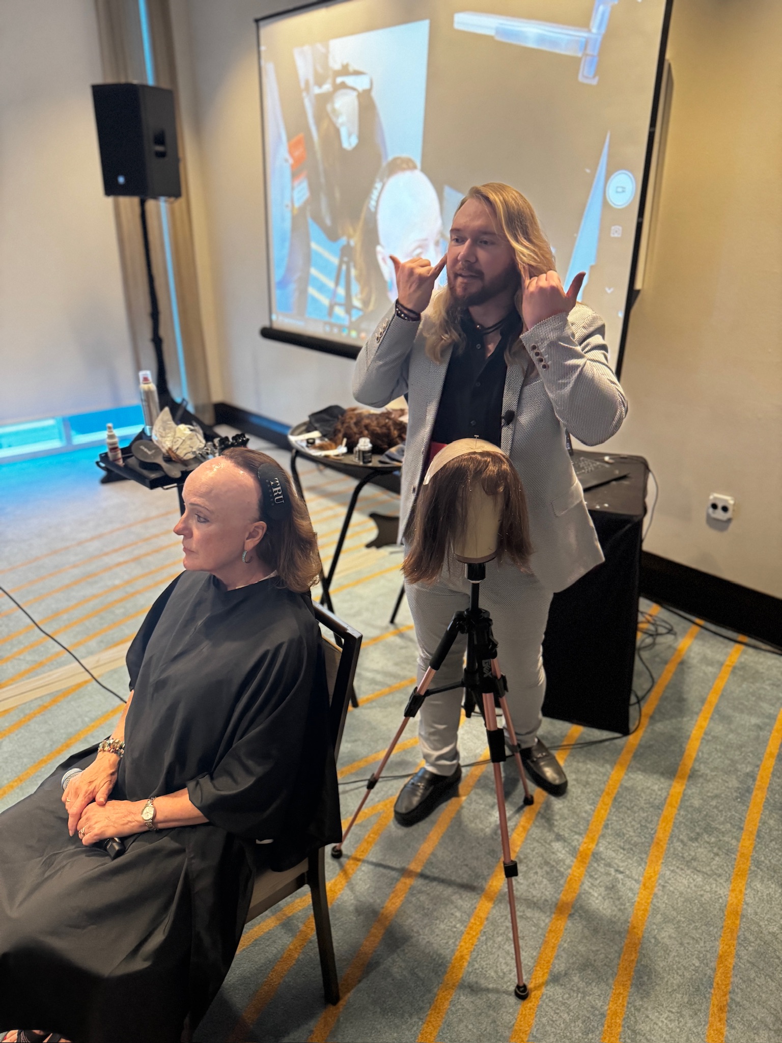 A woman with a shaved head and black outfit sits in a chair, while a man with long hair and beard stands behind her, gesturing with both hands. A mannequin head on a tripod is in front of him. A large screen and audio equipment are in the background.