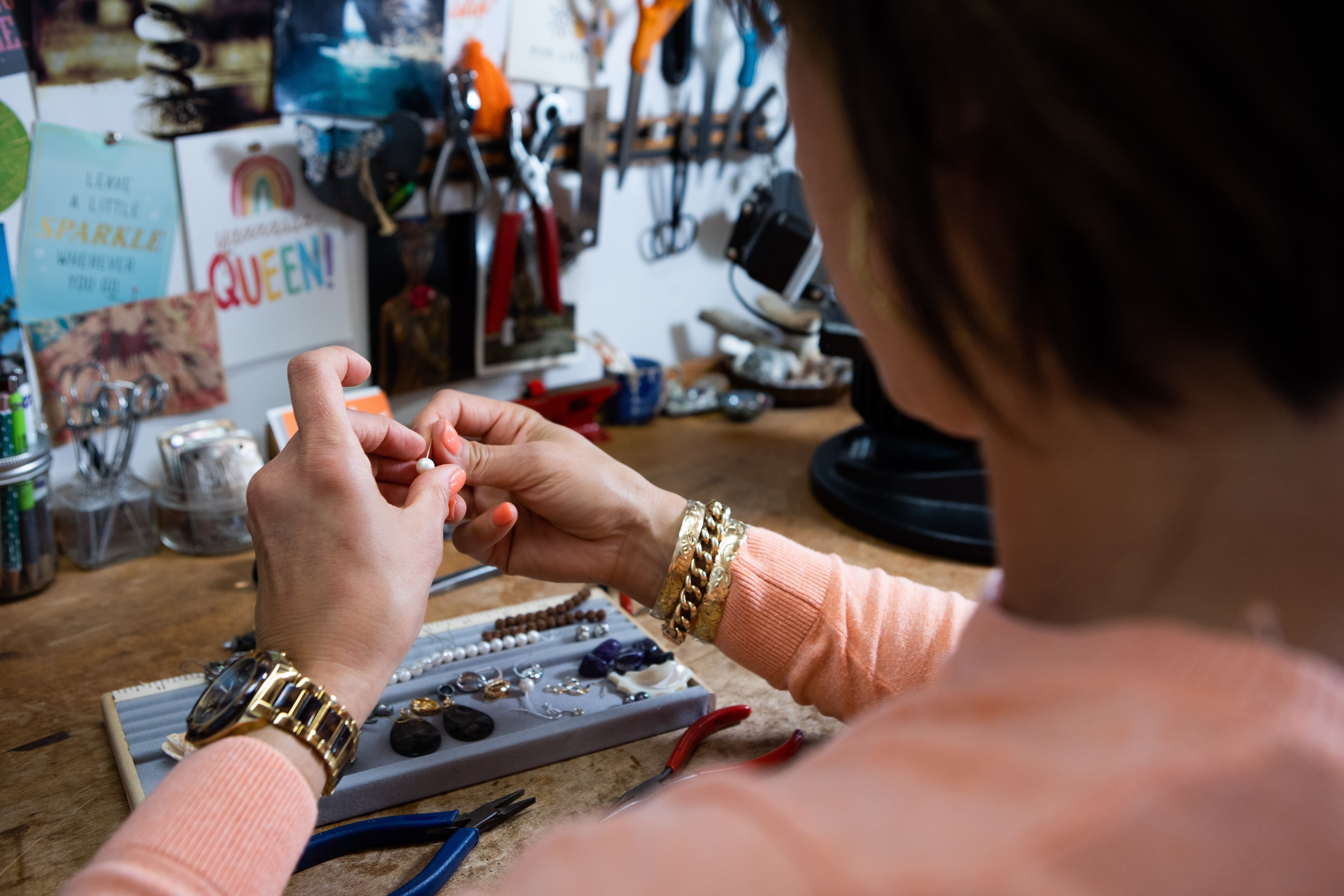Person working on jewelry at a cluttered workbench with tools and supplies, viewed from behind.