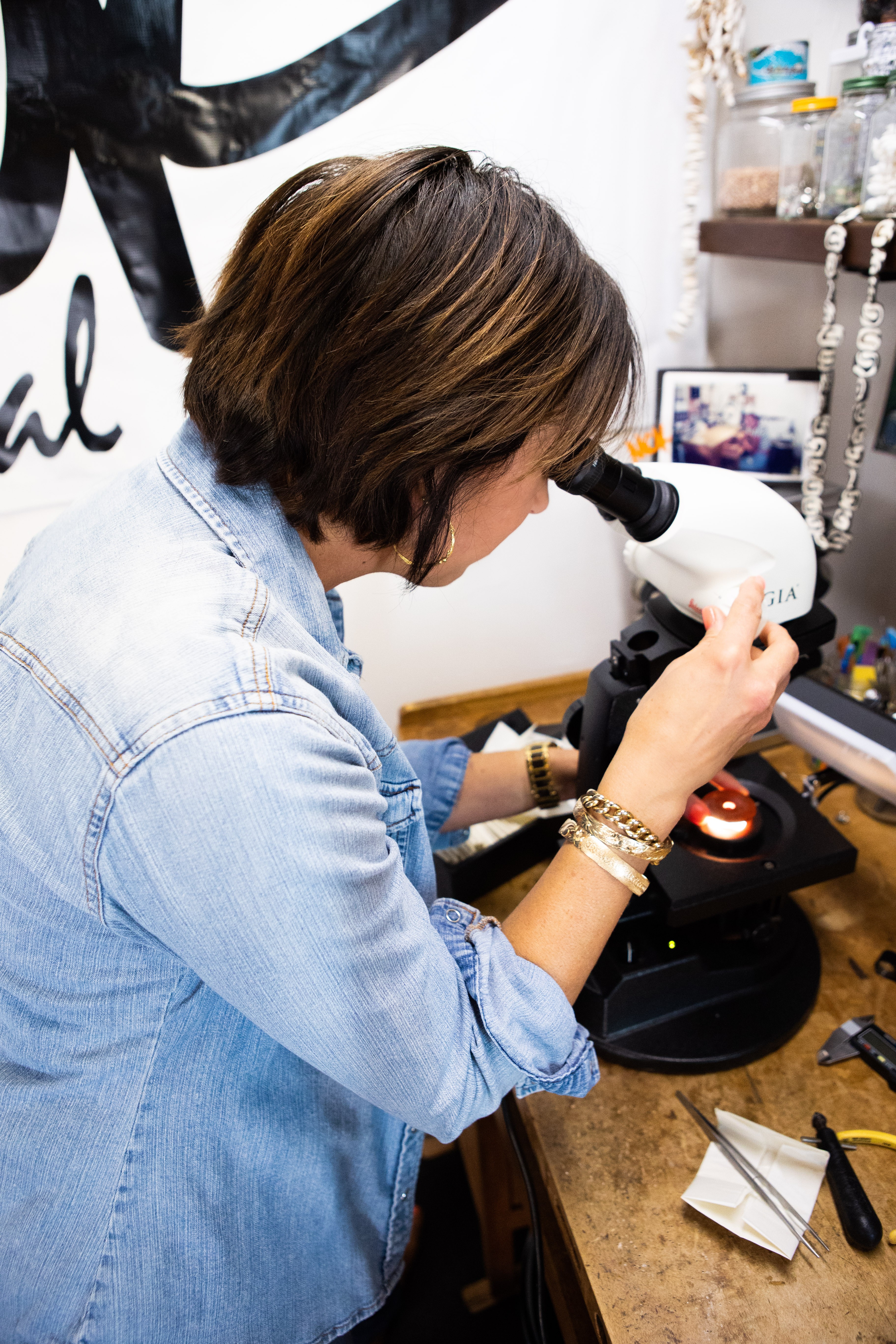 Woman looking into a microscope in a workspace with various items on the desk and shelves behind her.