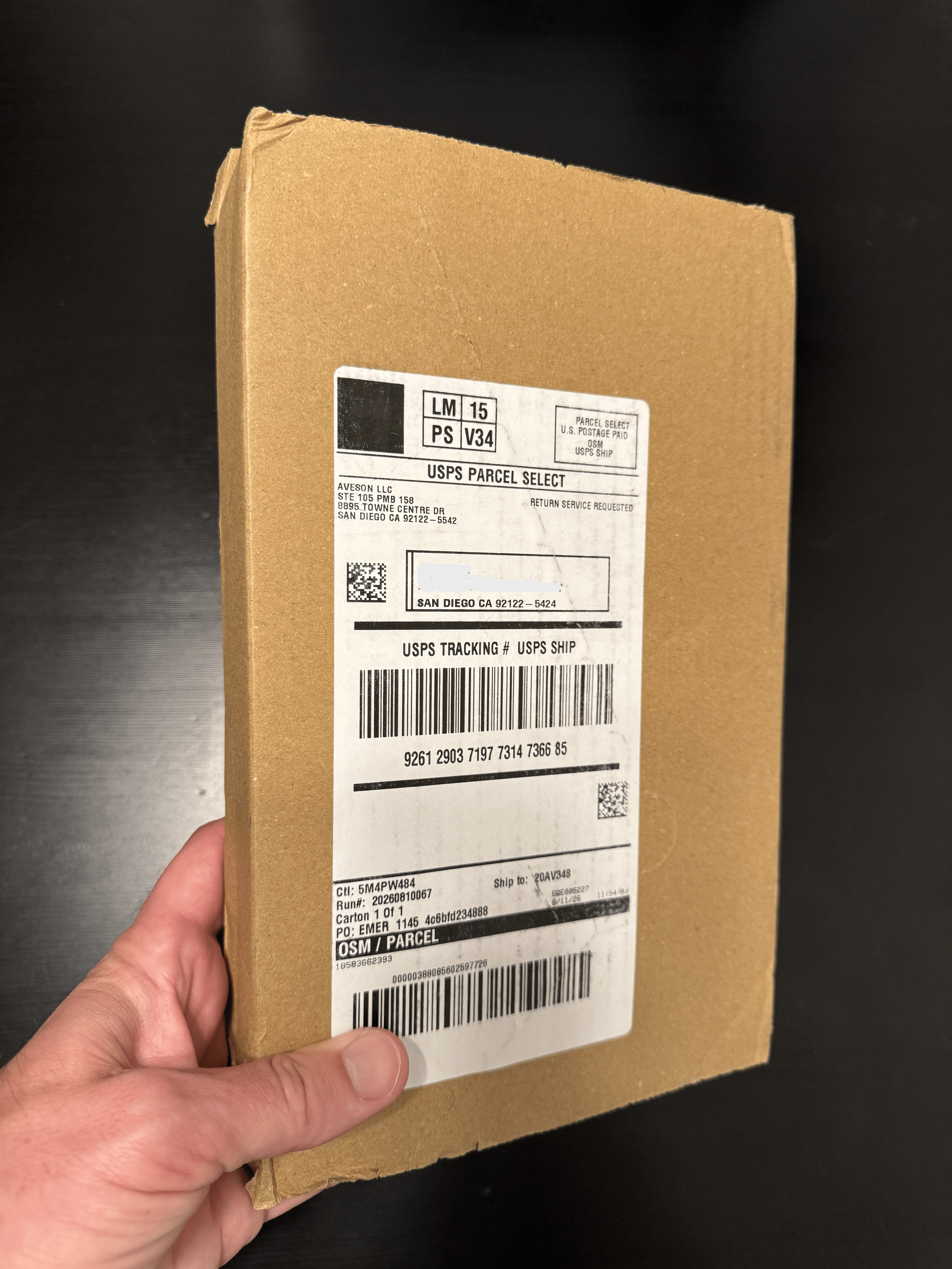 Hand holding a cardboard package with a shipping label, against a dark background.
