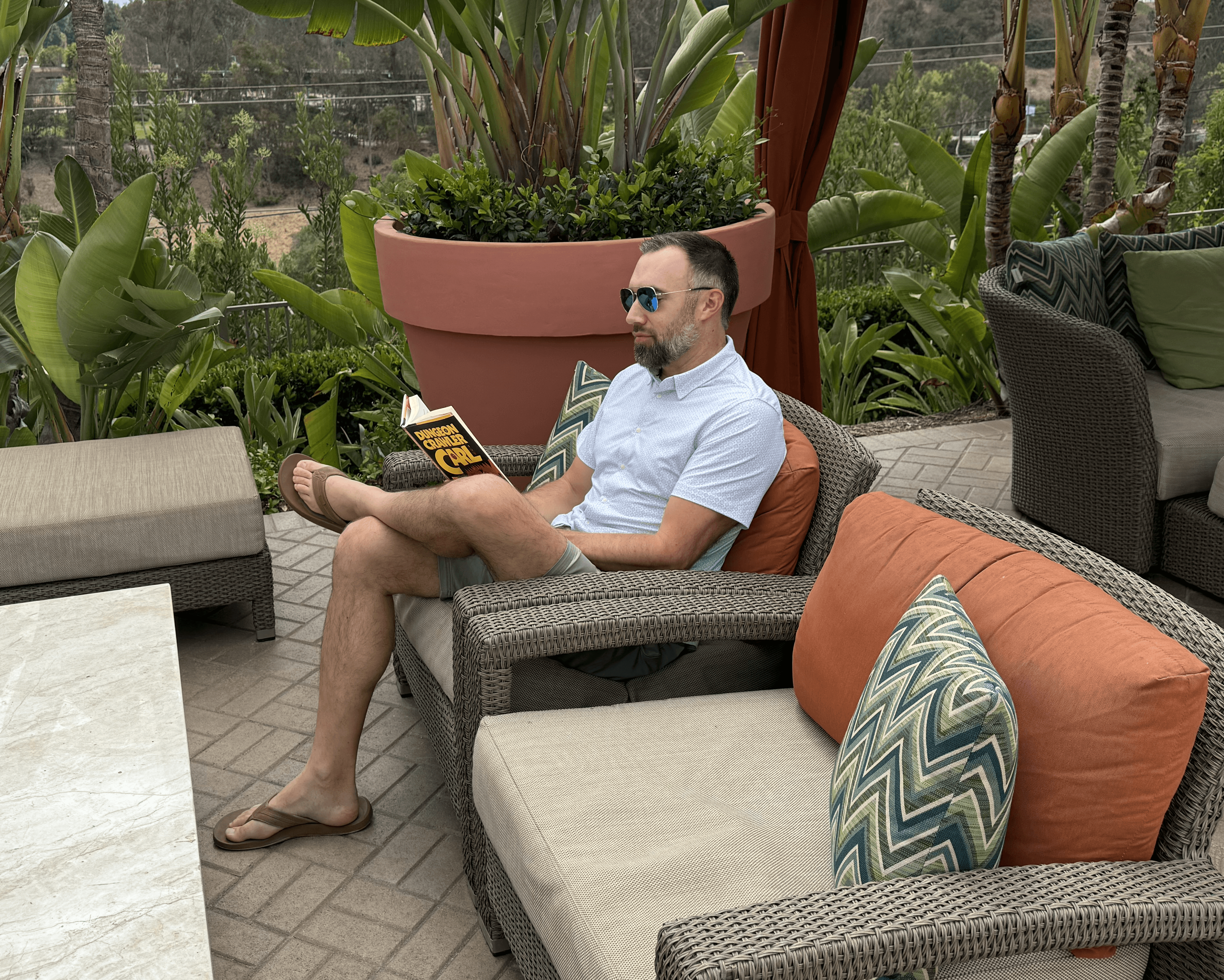 Man sitting outdoors on a wicker chair reading a book, surrounded by large potted plants and outdoor furniture.