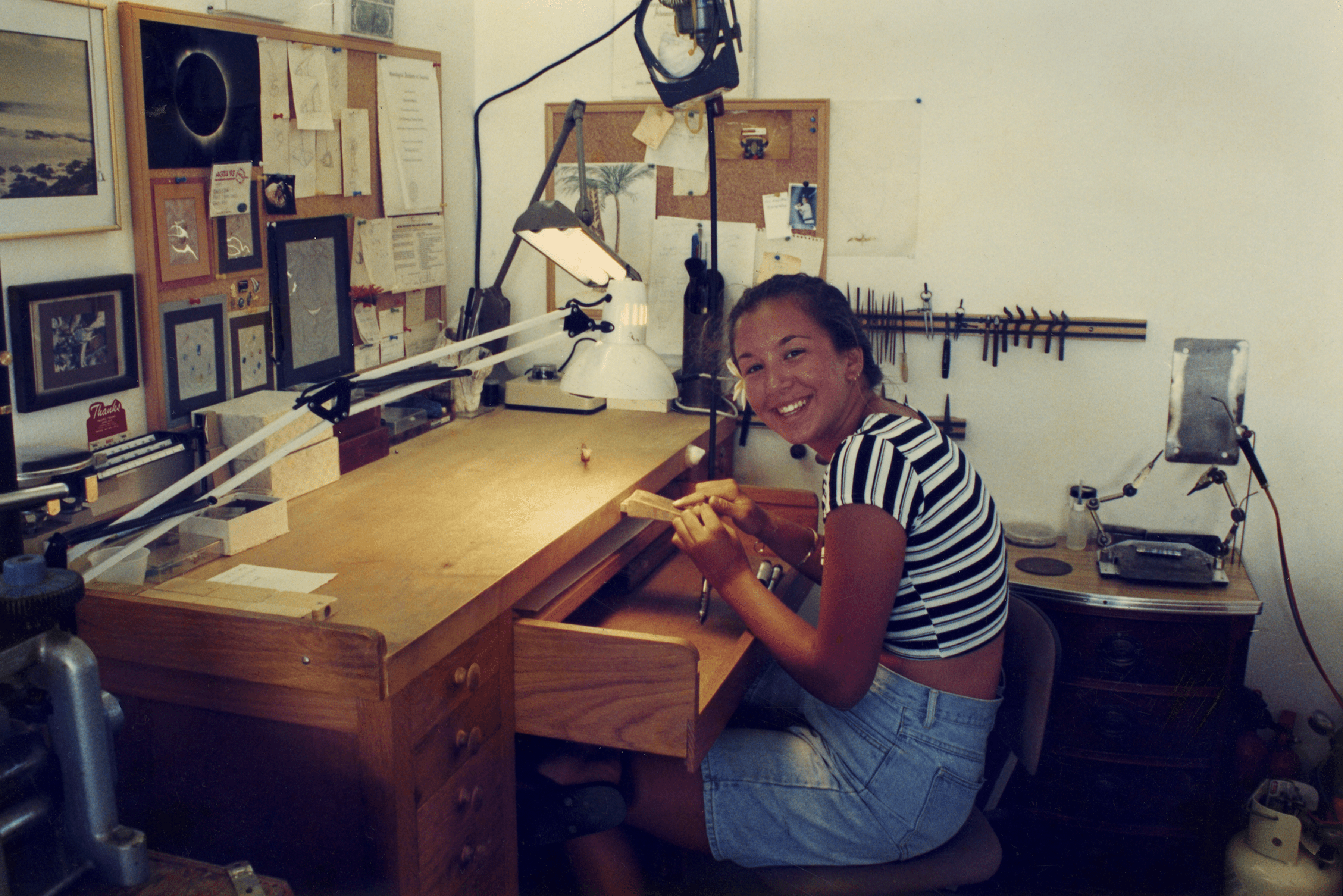 Young woman sitting at a workbench in a workshop, smiling and holding a small object, surrounded by tools and equipment.