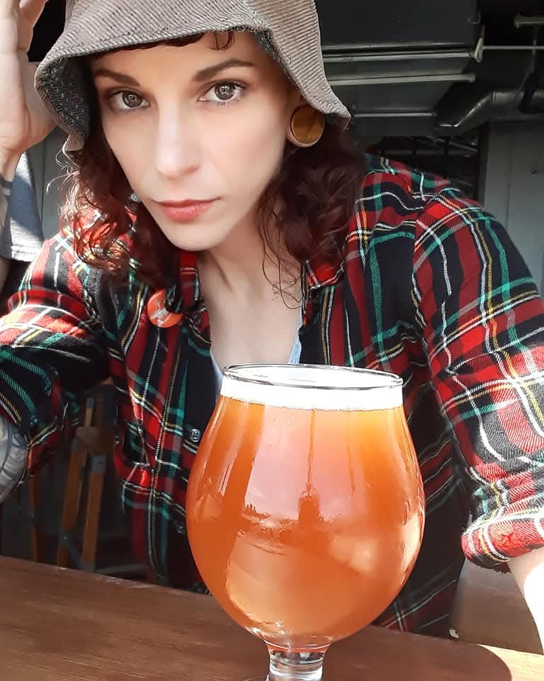 Woman with curly hair wearing a hat and plaid shirt, holding a glass of beer, sitting at a table.
