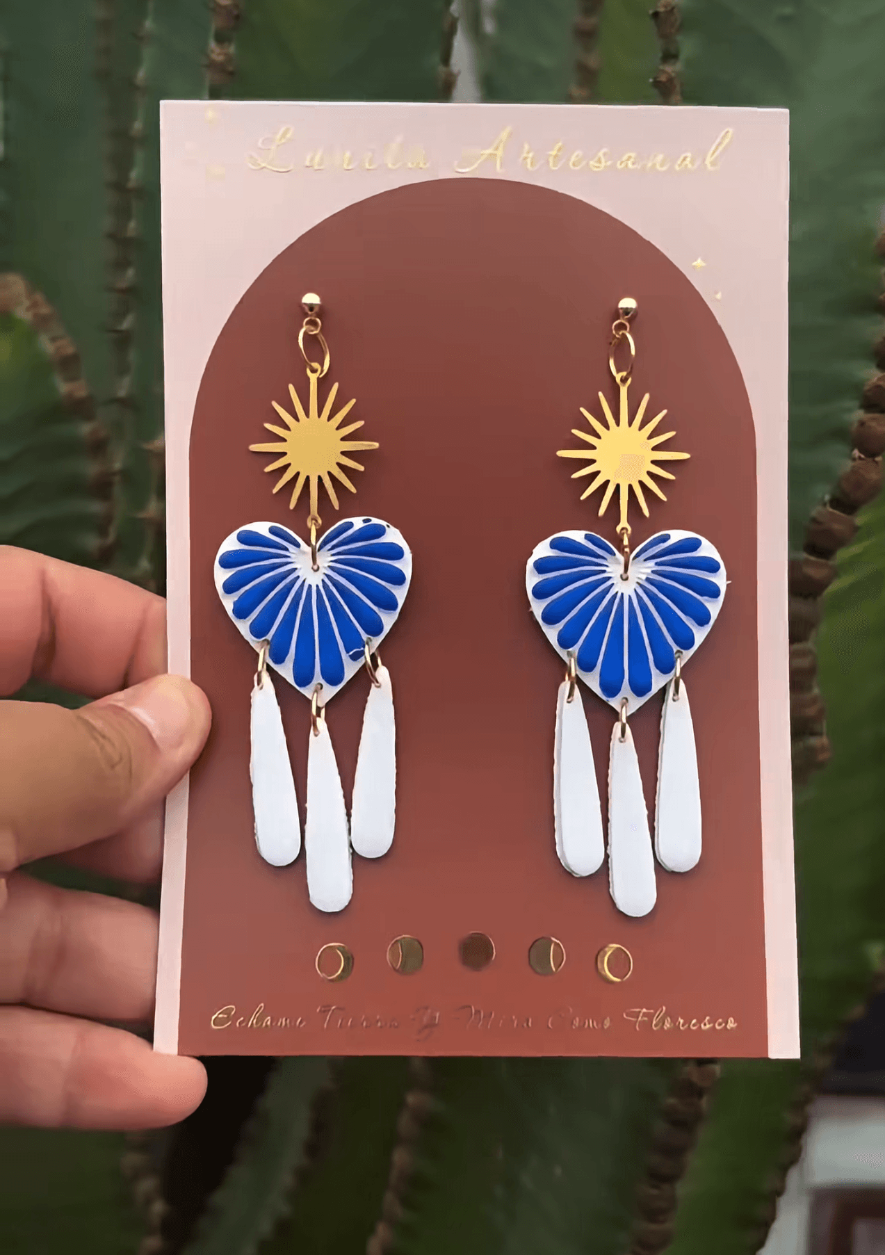 Hand holding colorful heart-shaped earrings with sun and feather designs on a display card, outdoors with green plants in background.