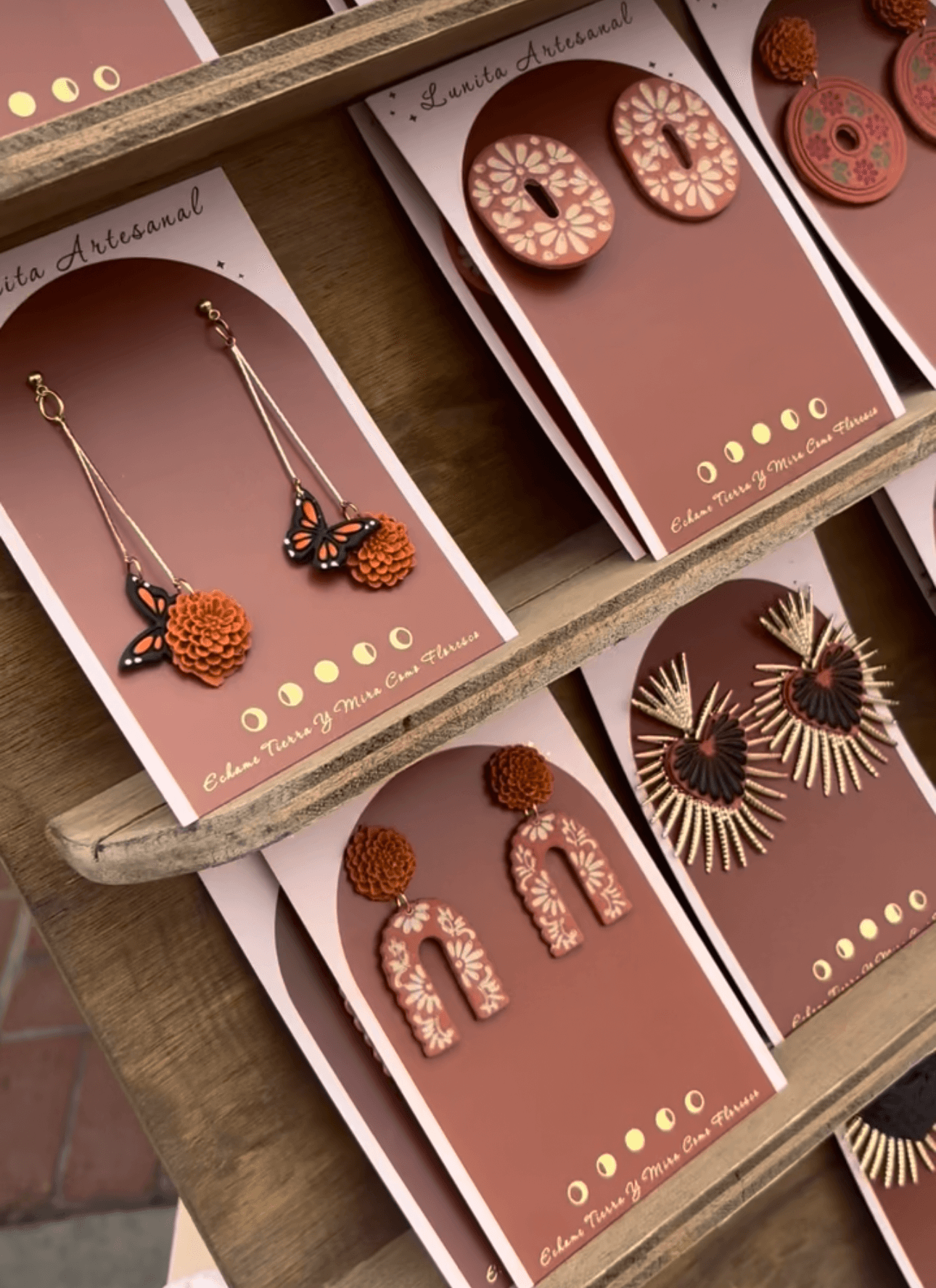 Display of colorful earrings with various shapes and patterns on pink cards, arranged on a wooden surface.