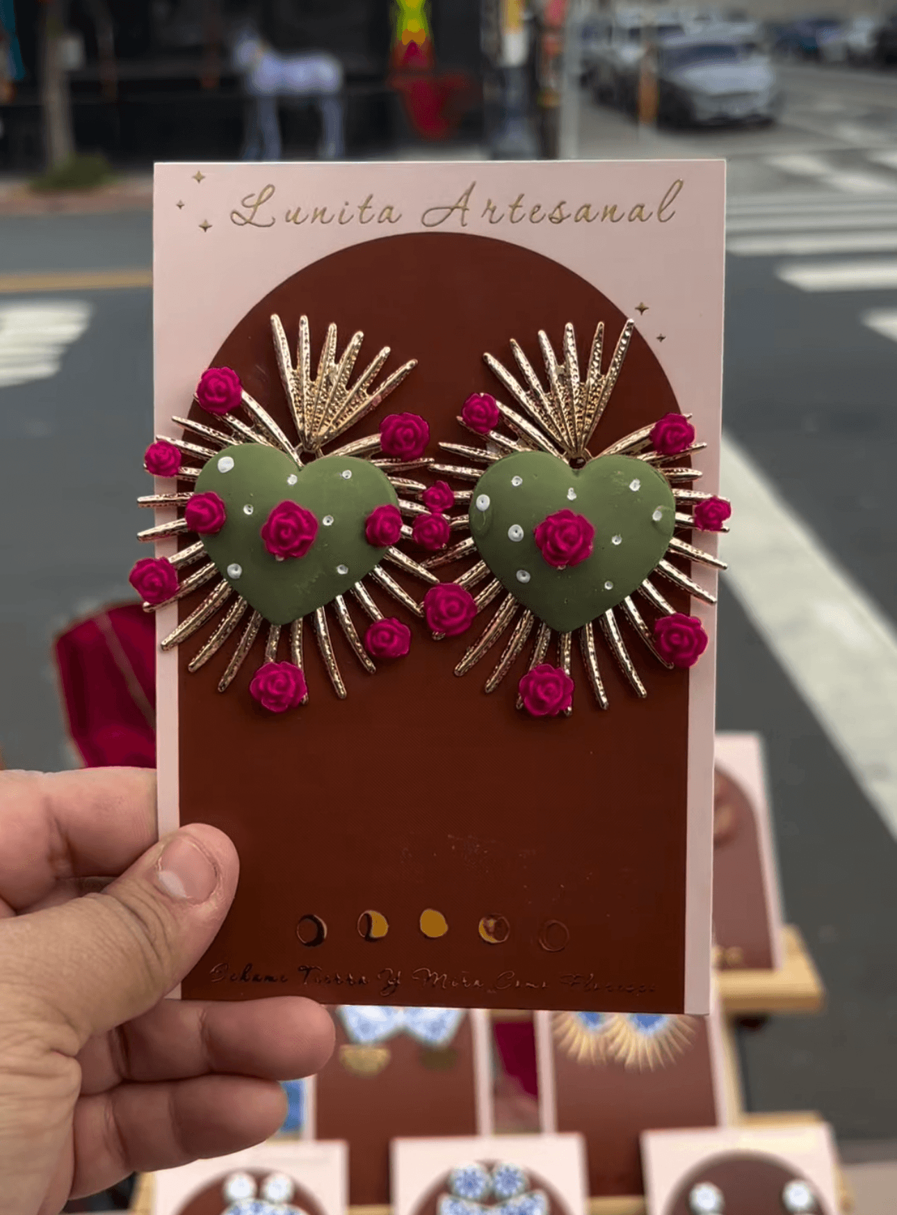 Hand holding decorative earrings with green hearts, pink accents, and gold rays, displayed on a pink card outdoors.