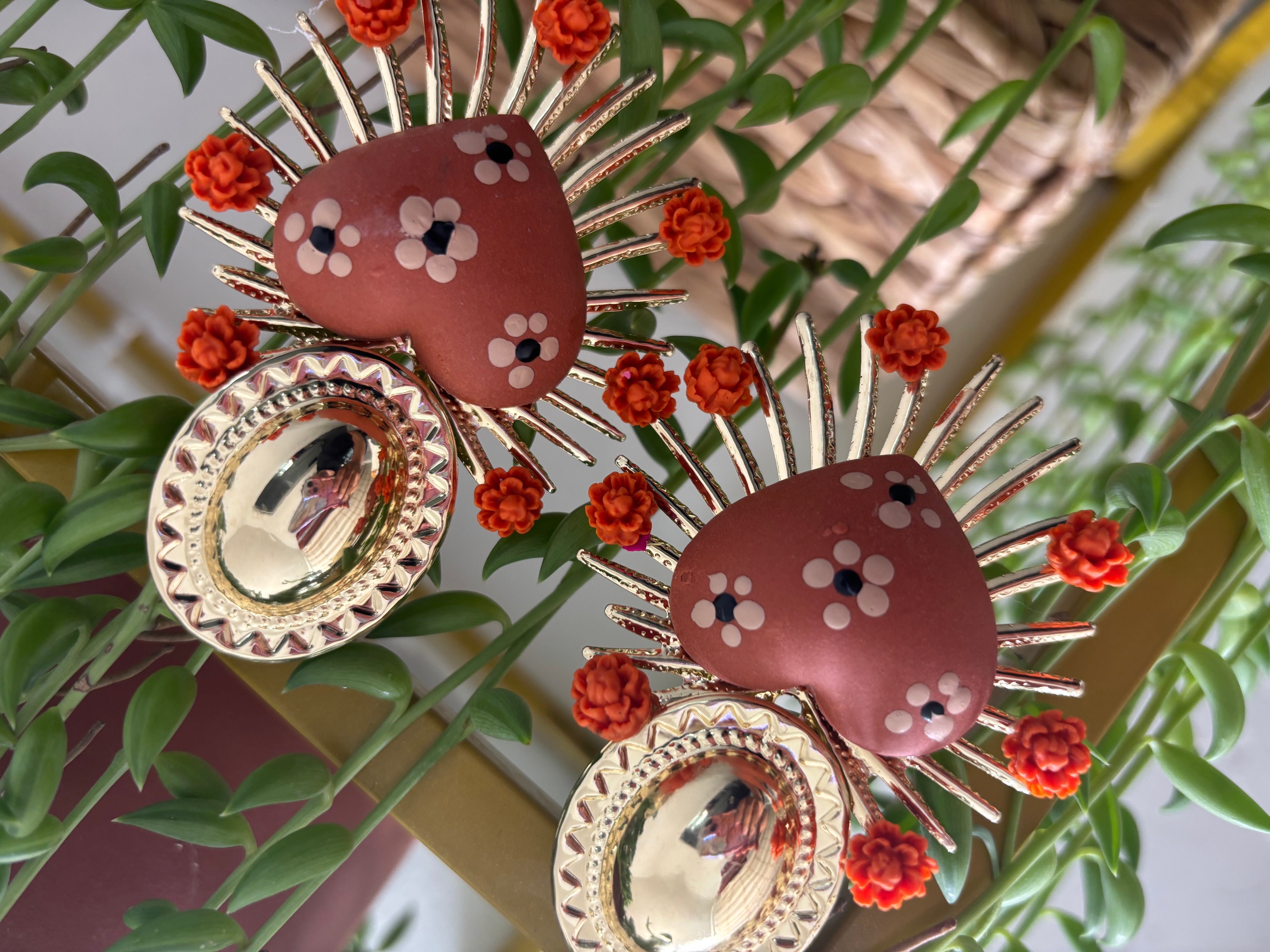 Decorative earrings with brown circular tops, white and black floral patterns, gold spikes, and small red flowers, hanging among green leaves.