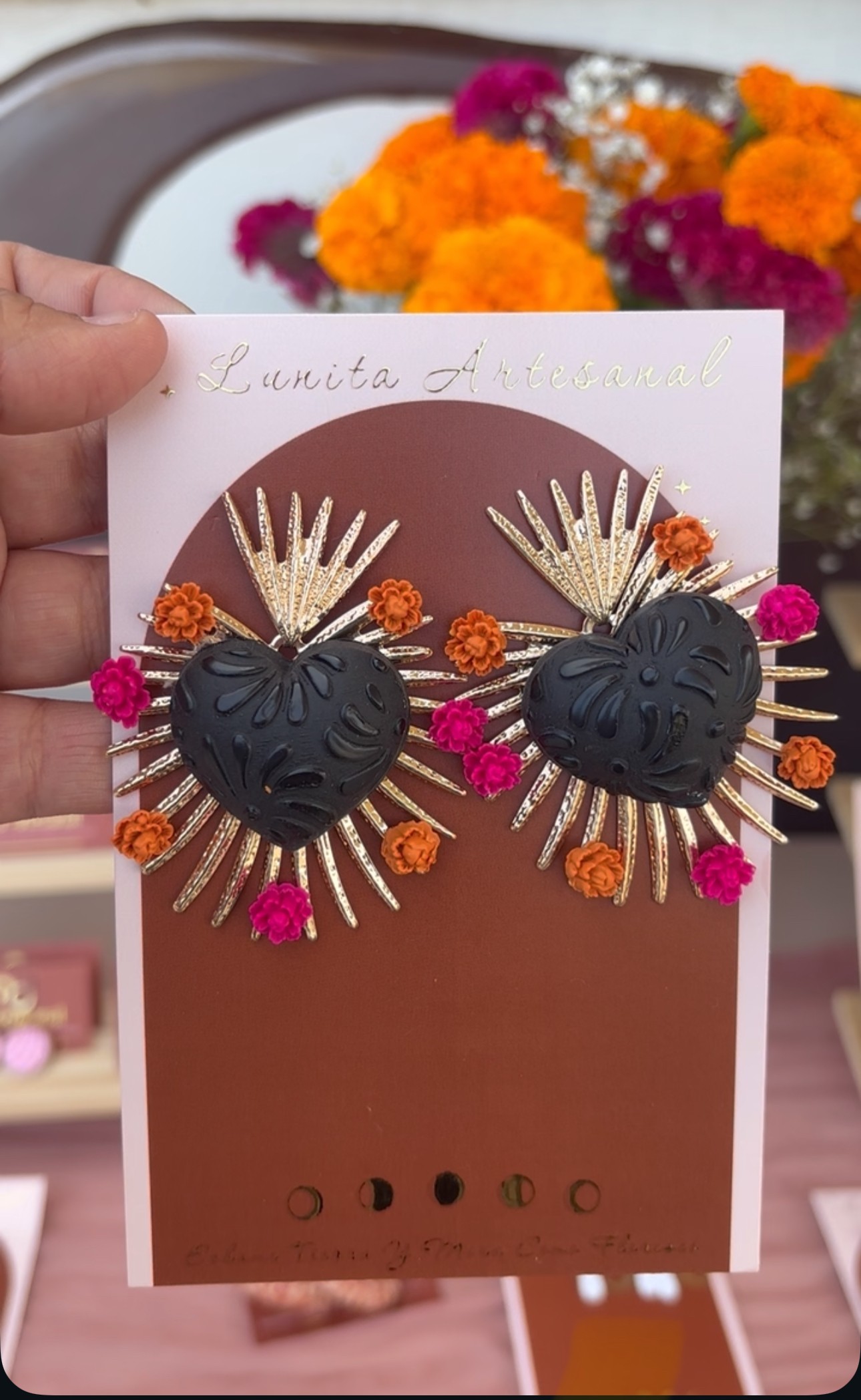 Hand holding a card with black heart-shaped earrings decorated with gold and pink accents, with colorful flowers in the background.