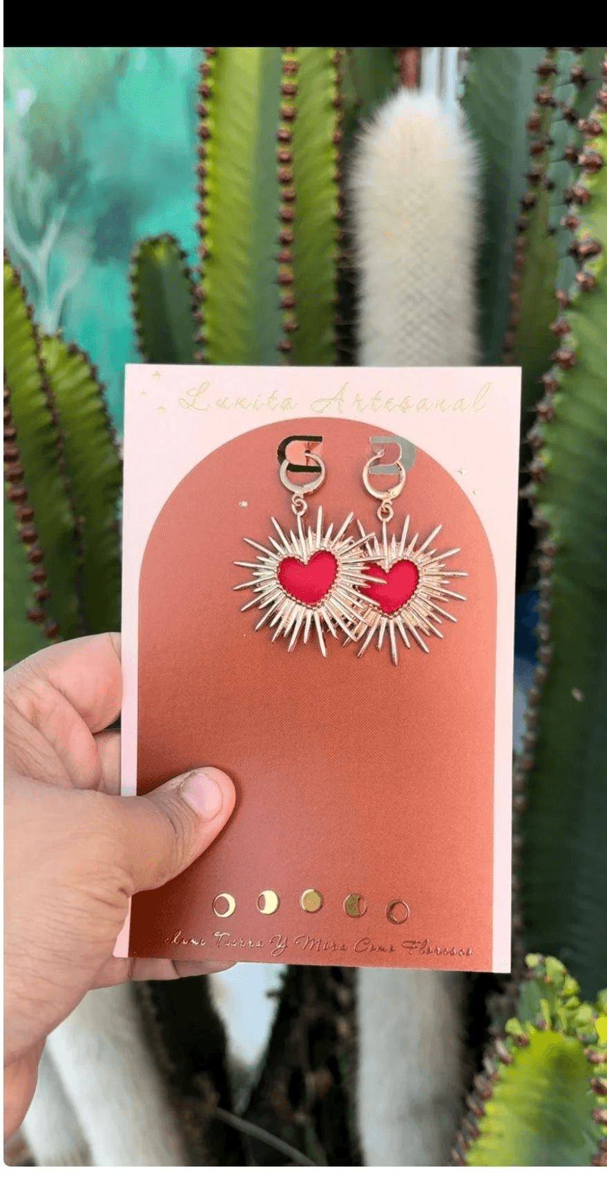Hand holding a card with earrings featuring hearts and sunburst designs, cactus plants in background.