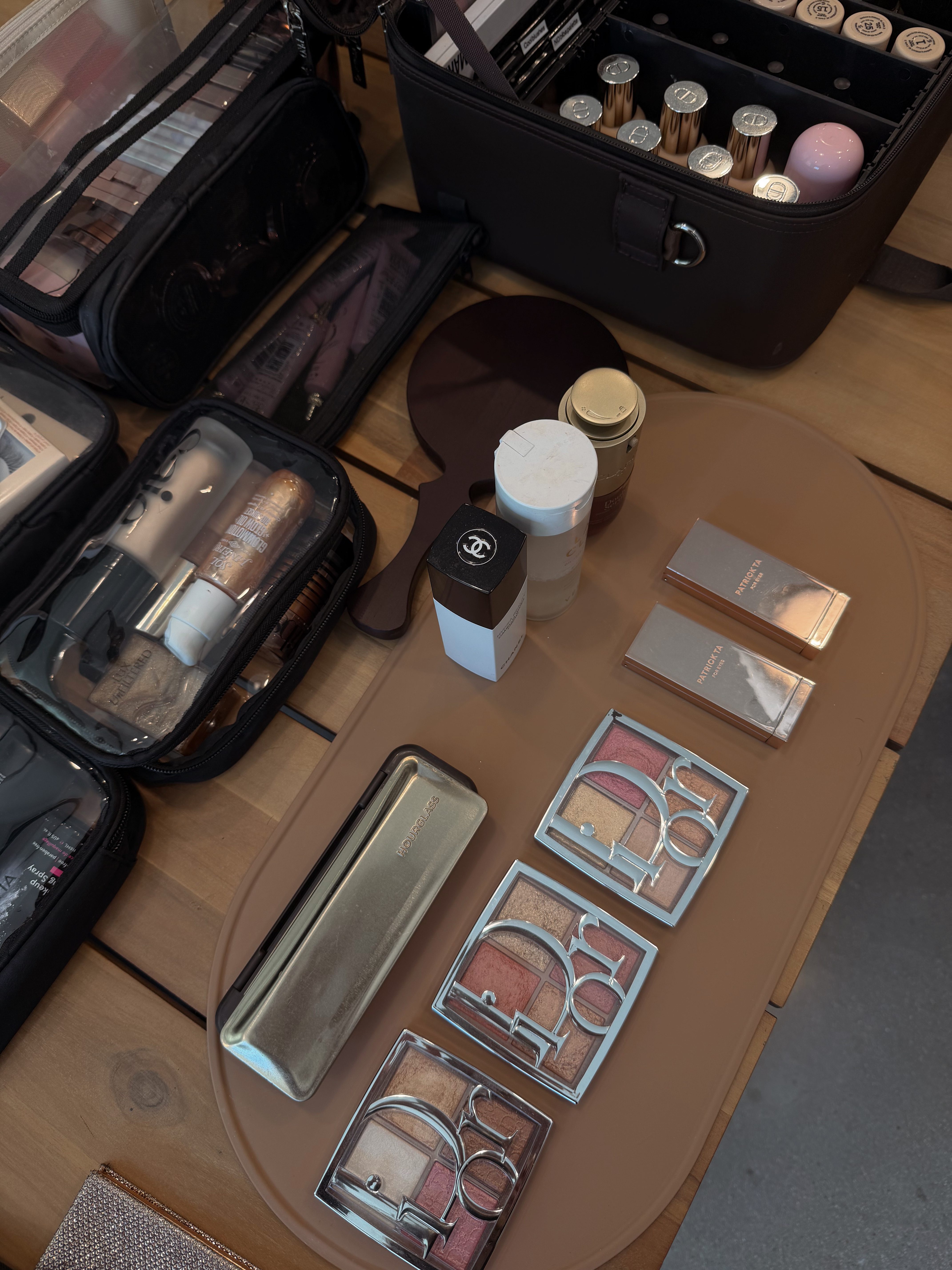 Makeup products and tools on a wooden table, including palettes, brushes, and containers, with a black case nearby.