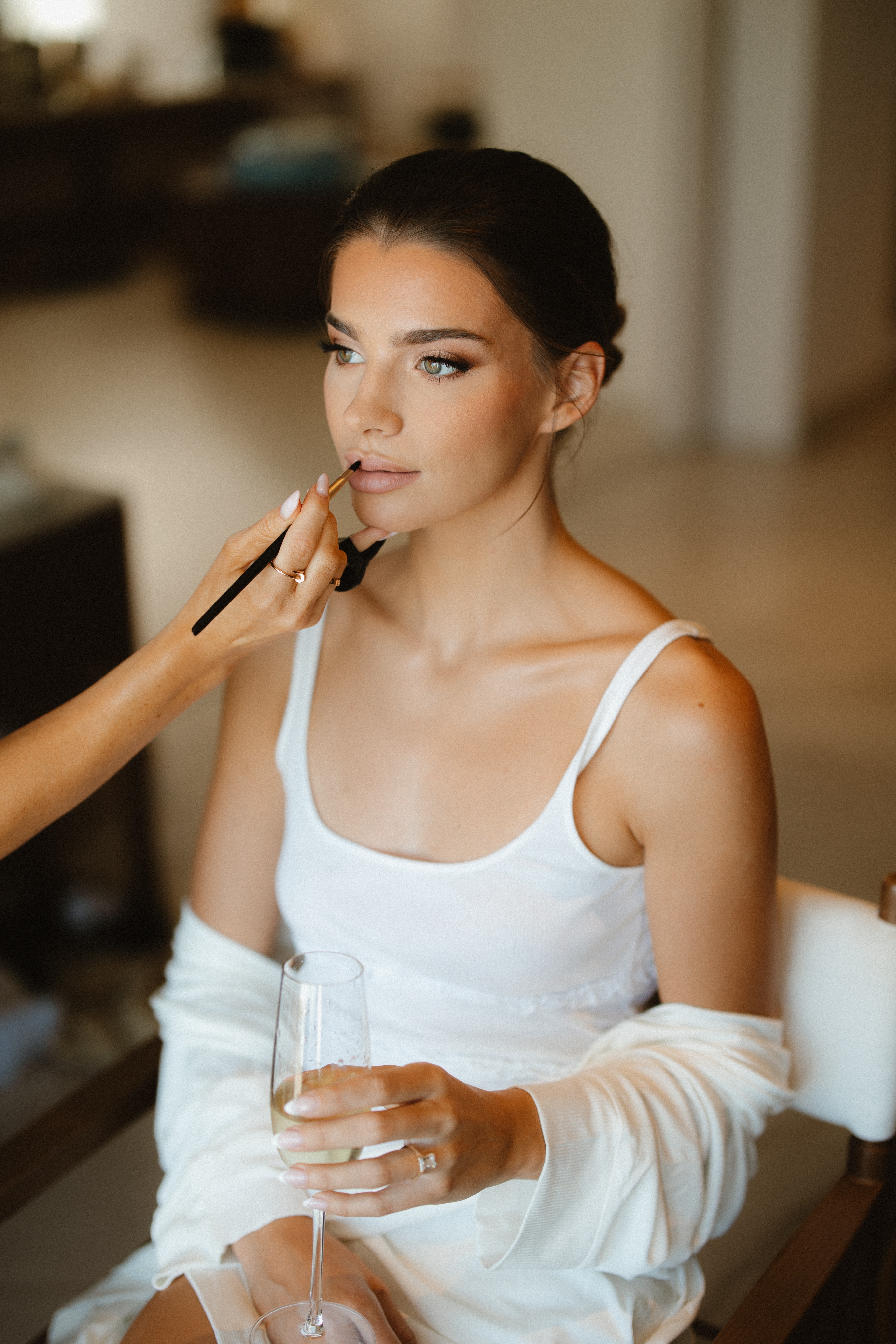 Woman with dark hair in a white top getting makeup applied, holding a glass, in an indoor setting.