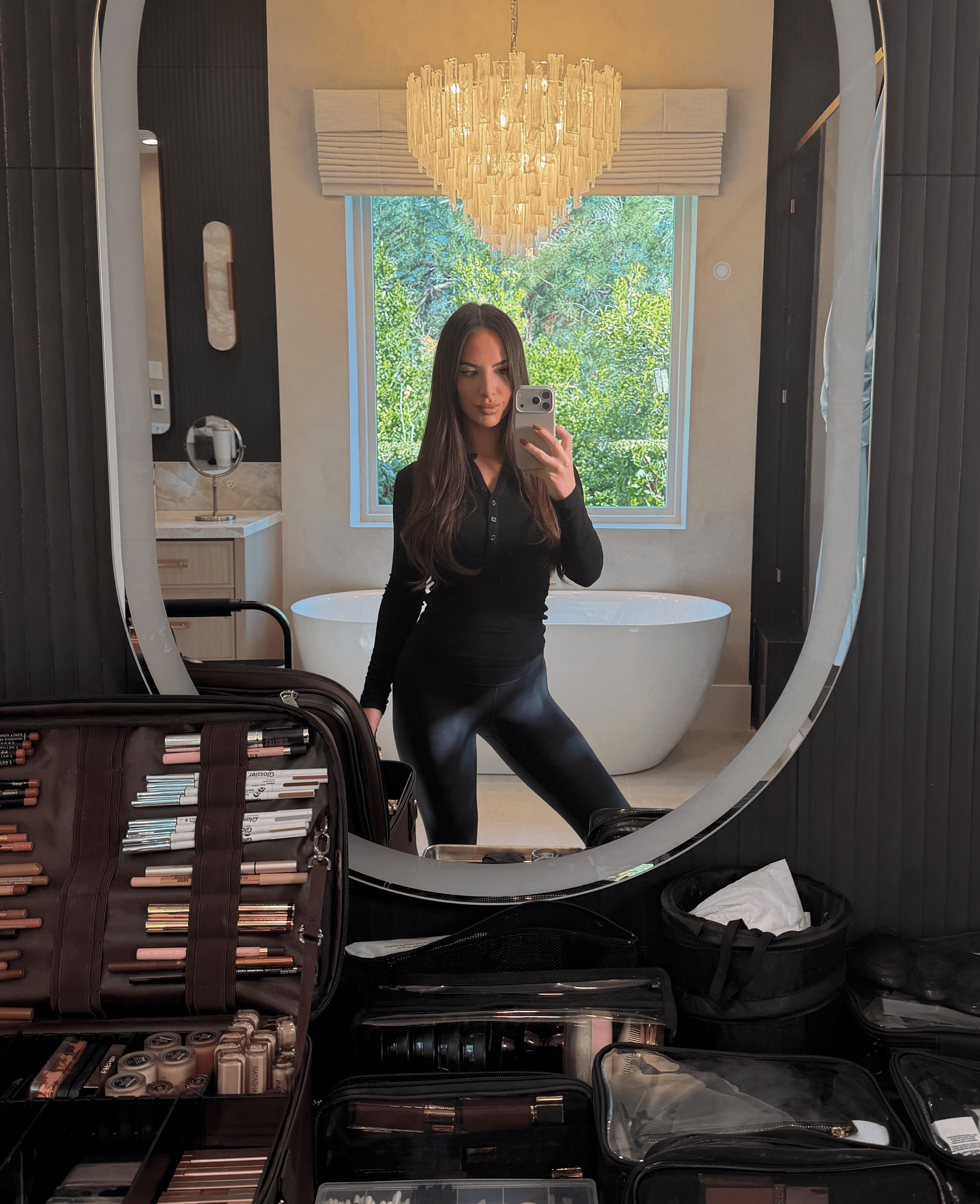 Woman taking a mirror selfie in a bathroom with a chandelier and bathtub, makeup products in foreground.