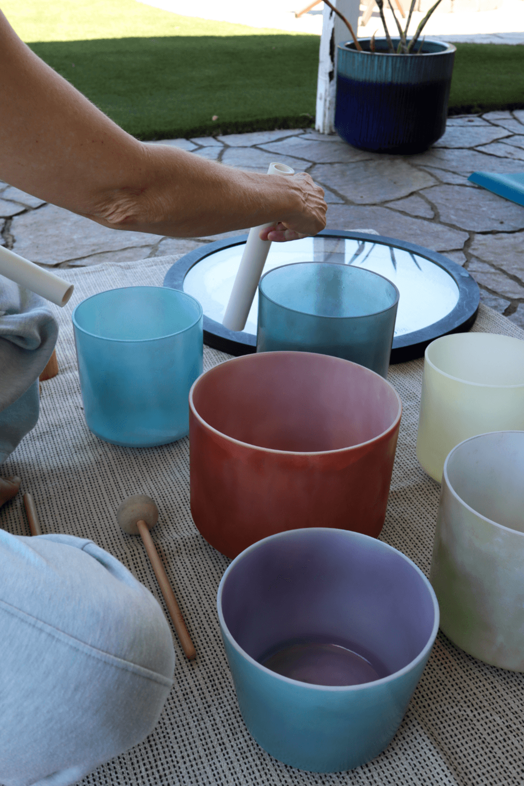 Person's arm reaching for a glass on a table with colorful cups, outdoor setting, potted plant in background.