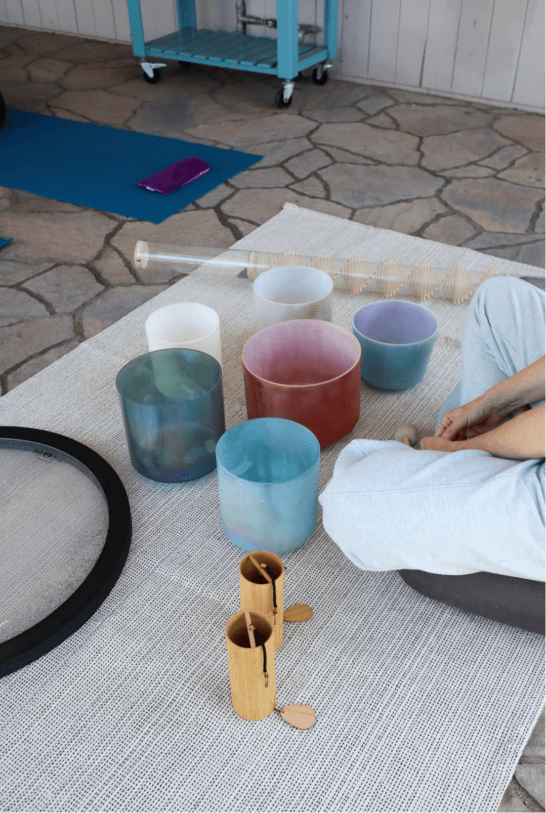 Person sitting on a rug with colorful glass cups, a white bowl, and wooden objects nearby, outdoors on stone pavement.