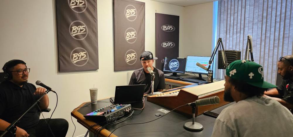 Four people in a radio studio with microphones, laptops, and sound equipment, walls with B95 logos, and a window with blinds.