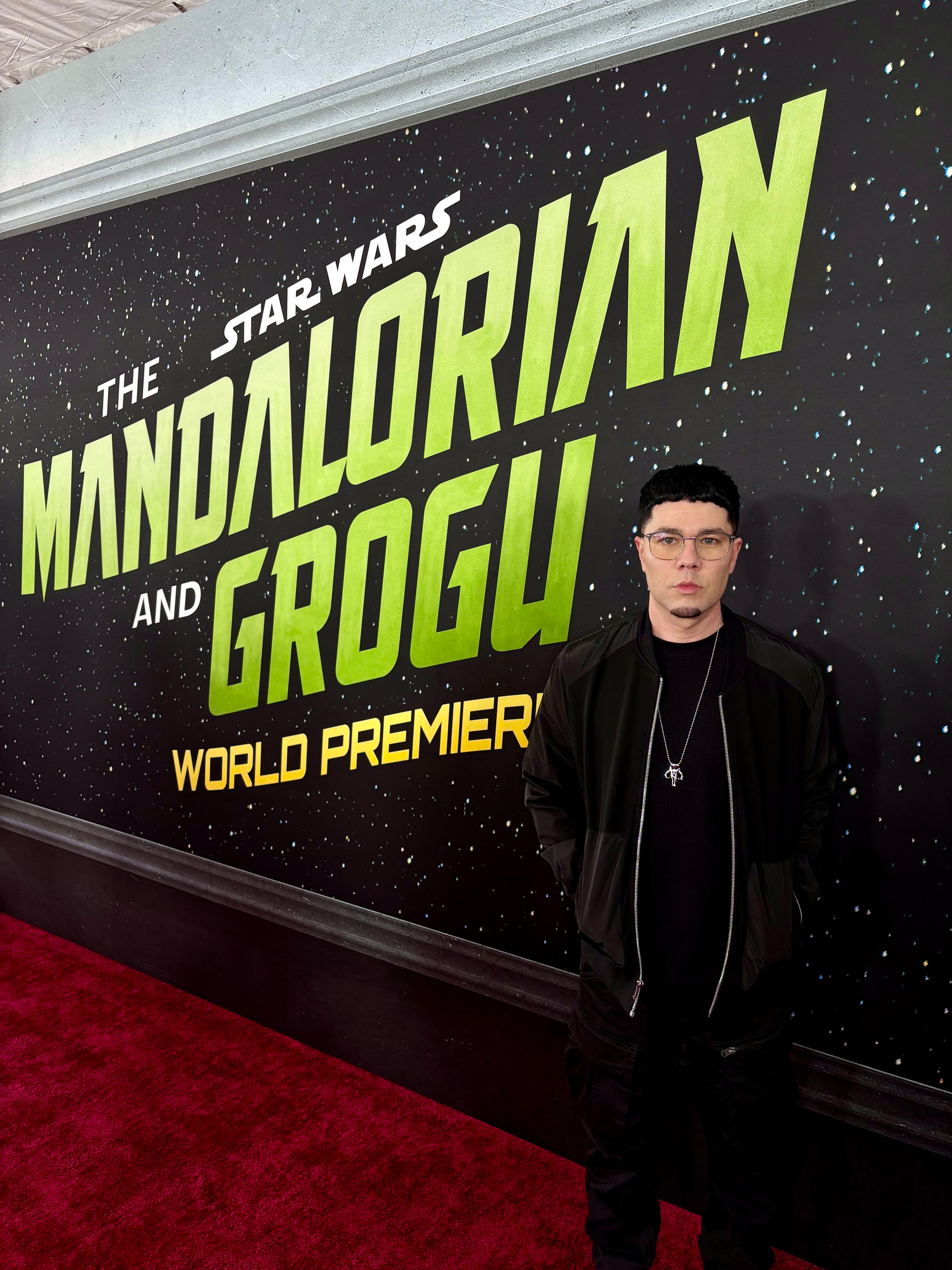 Young man with glasses and dark clothing standing in front of a Star Wars Mandalorian and Grogu poster, on a red carpet.