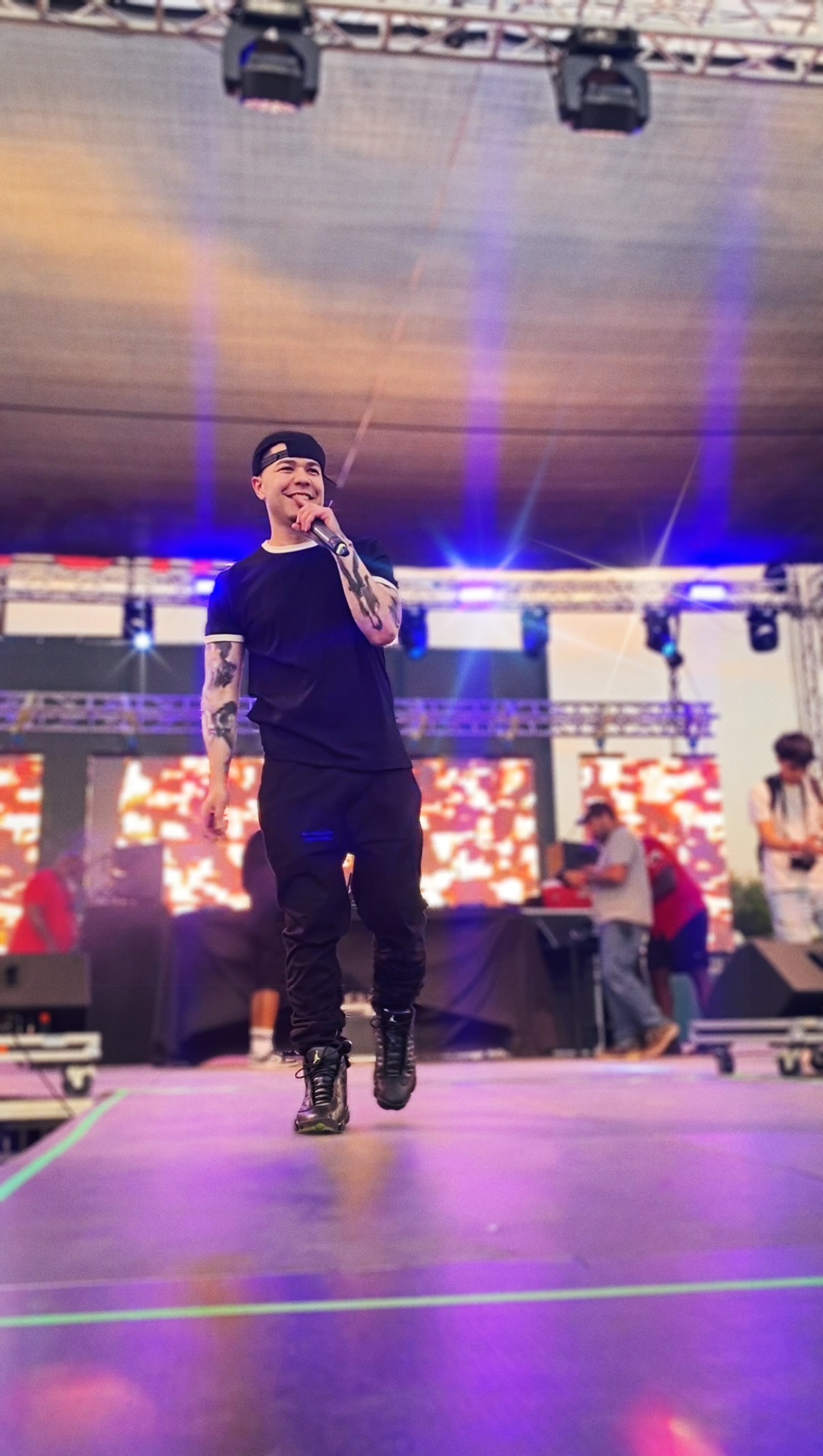 Performer on stage holding microphone, wearing black outfit, with colorful LED screens behind, stage lights overhead, and people in background.