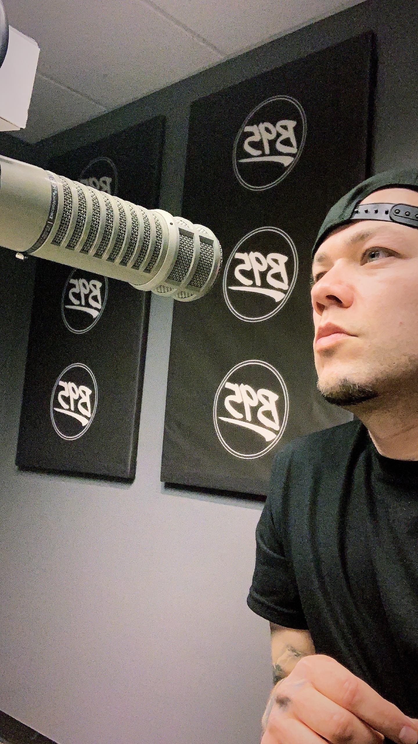 Person wearing a black cap and shirt, looking to the right, in a room with black wall panels and circular logos.