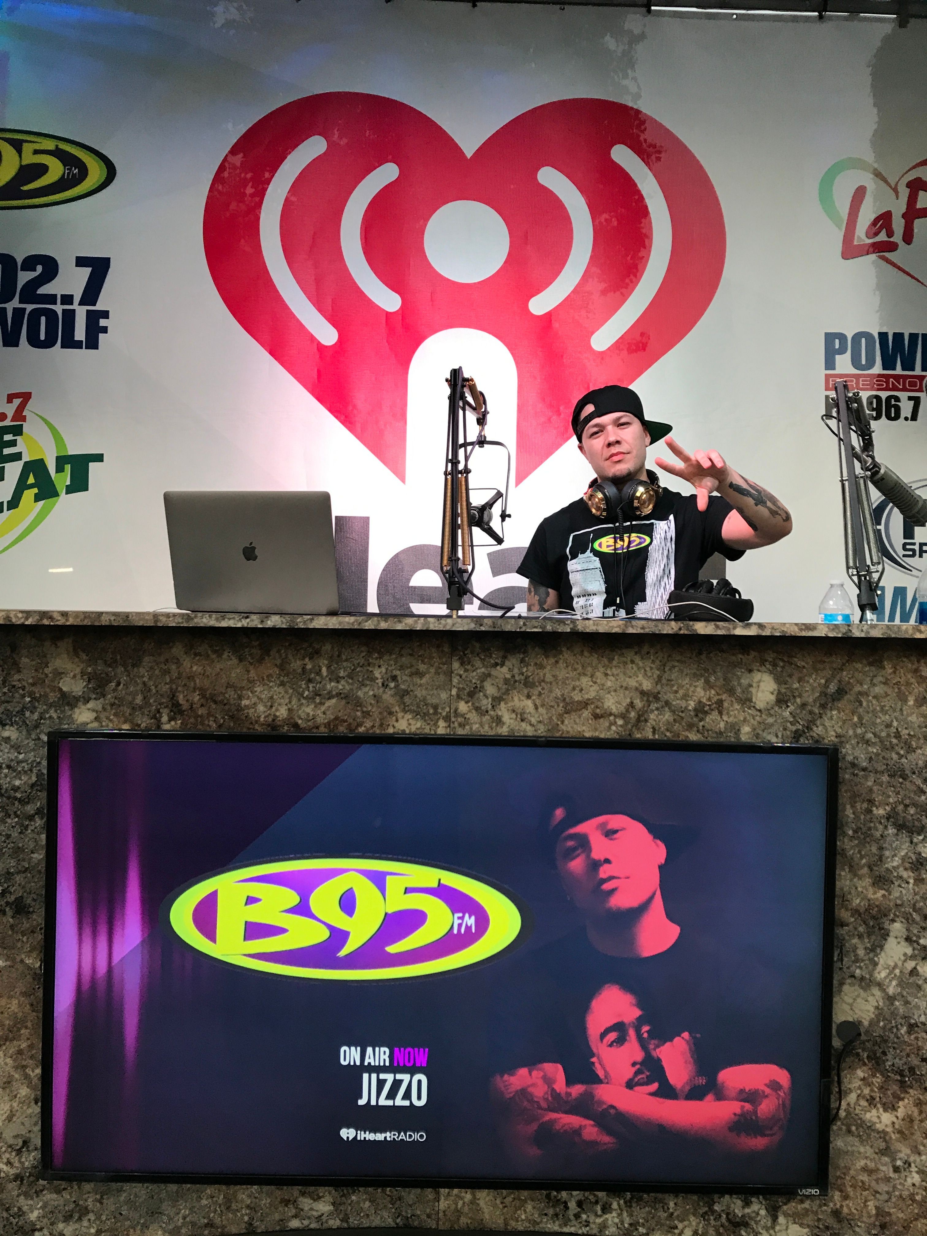 Person sitting at a table with a microphone, large red heart logo on the wall behind, and various logos around.
