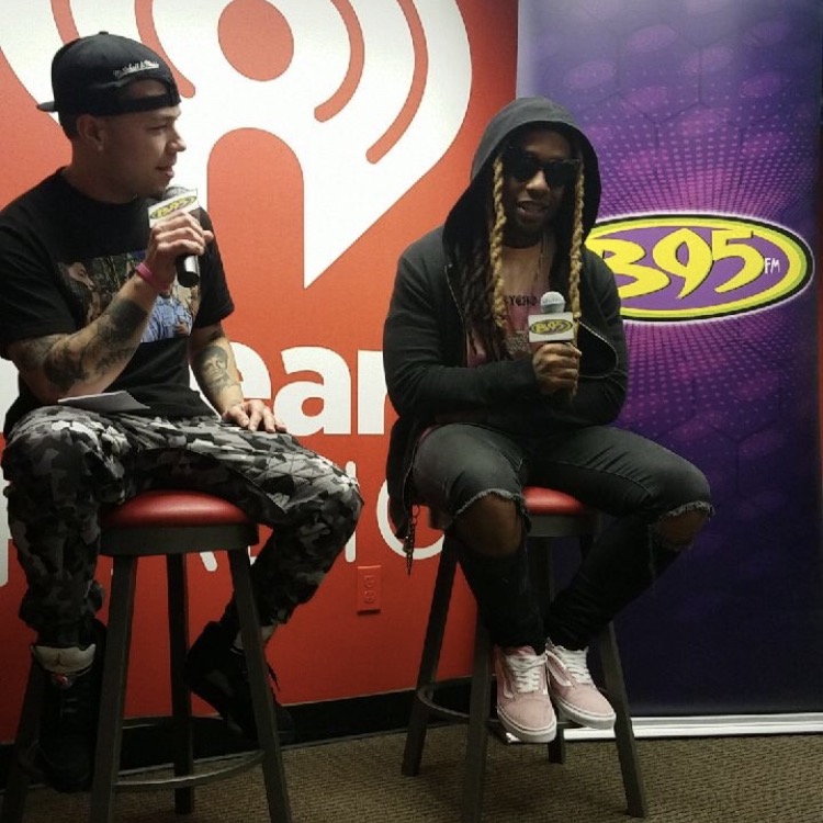Two people sitting on stools holding microphones, one wearing sunglasses and a hoodie, the other in a cap and patterned pants.