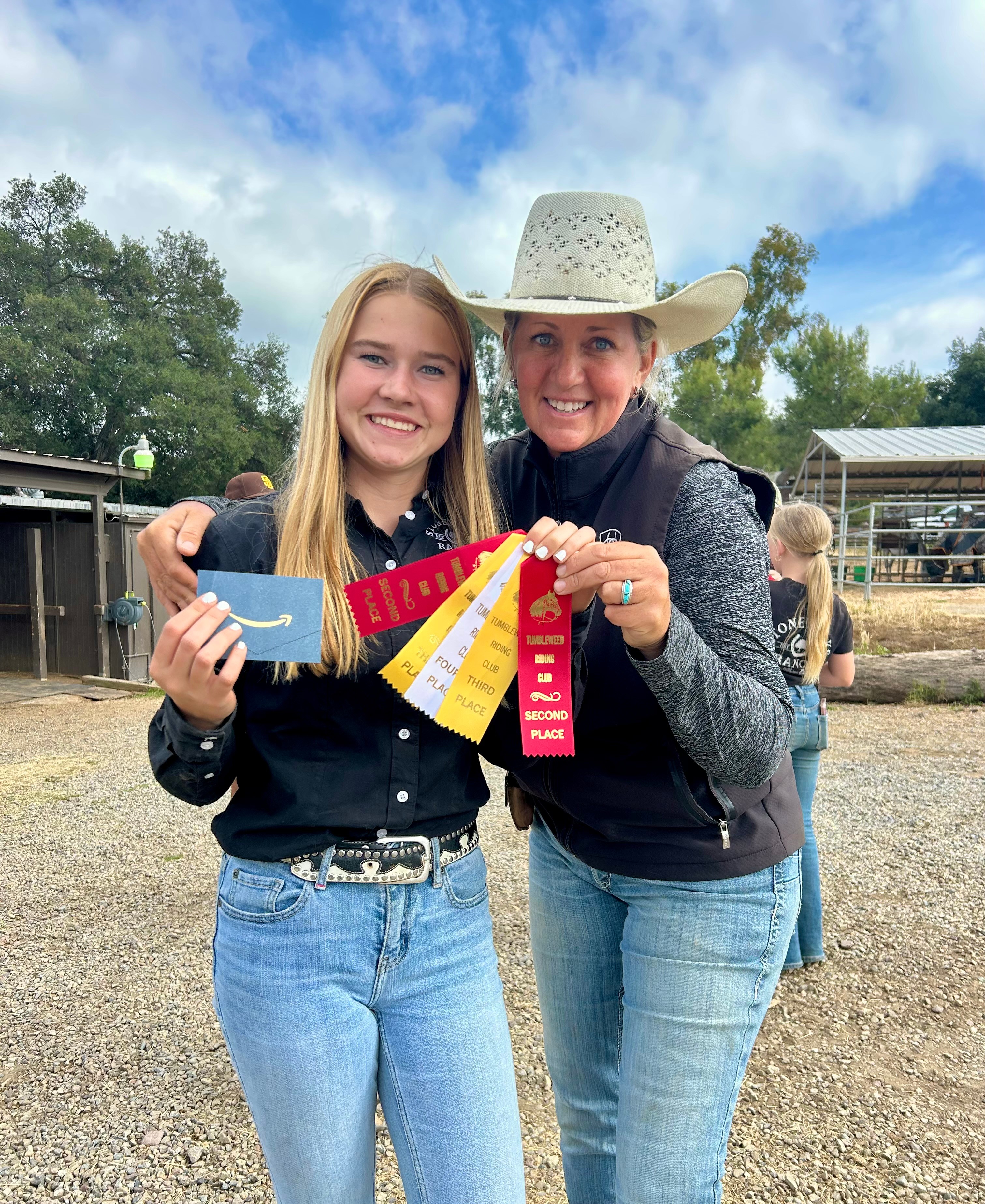 Two women smiling outdoors, one holding colorful ribbons, with trees and a barn in the background.