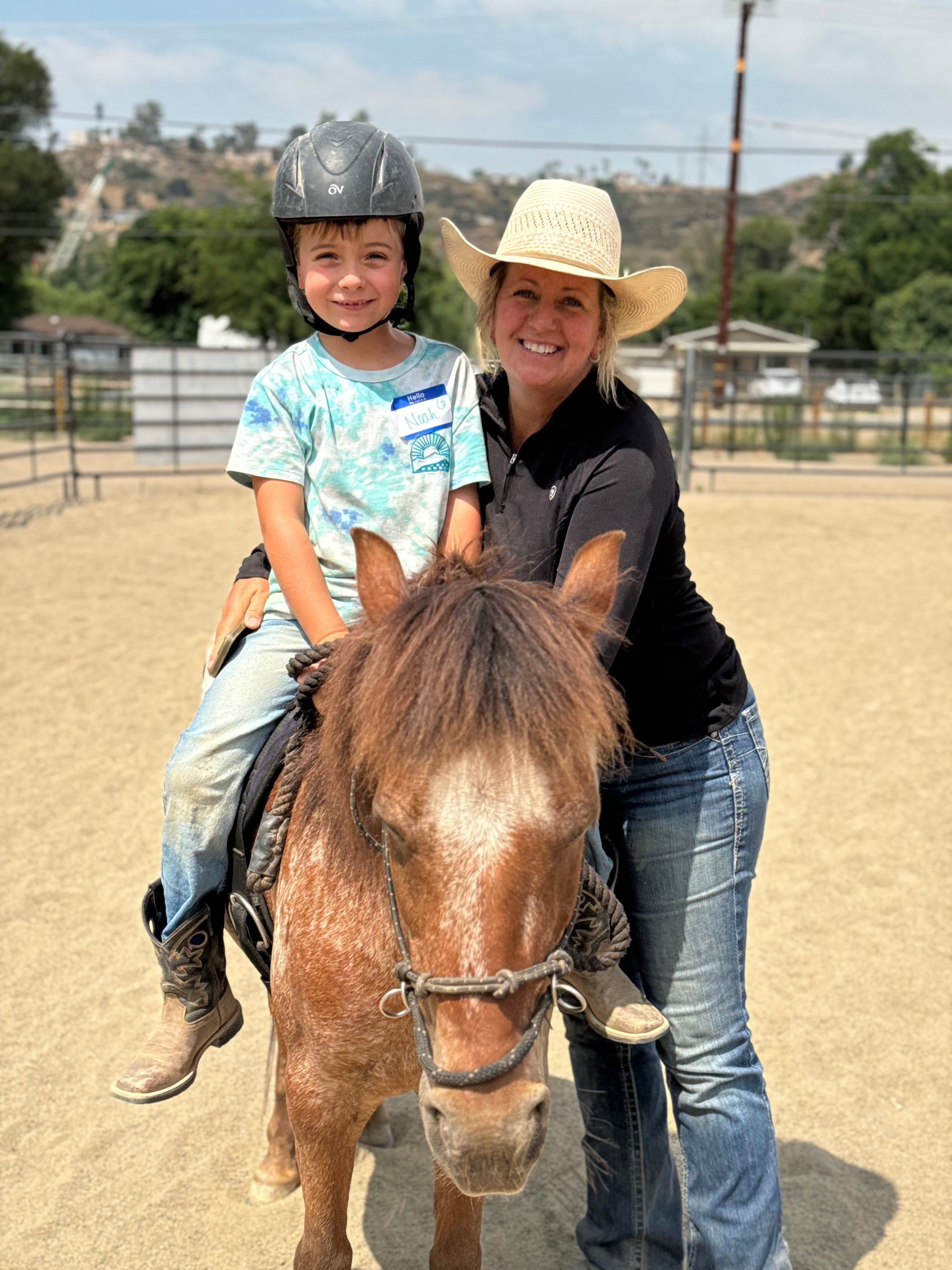 Child wearing a helmet riding a small horse, with a woman in a hat standing beside them outdoors.