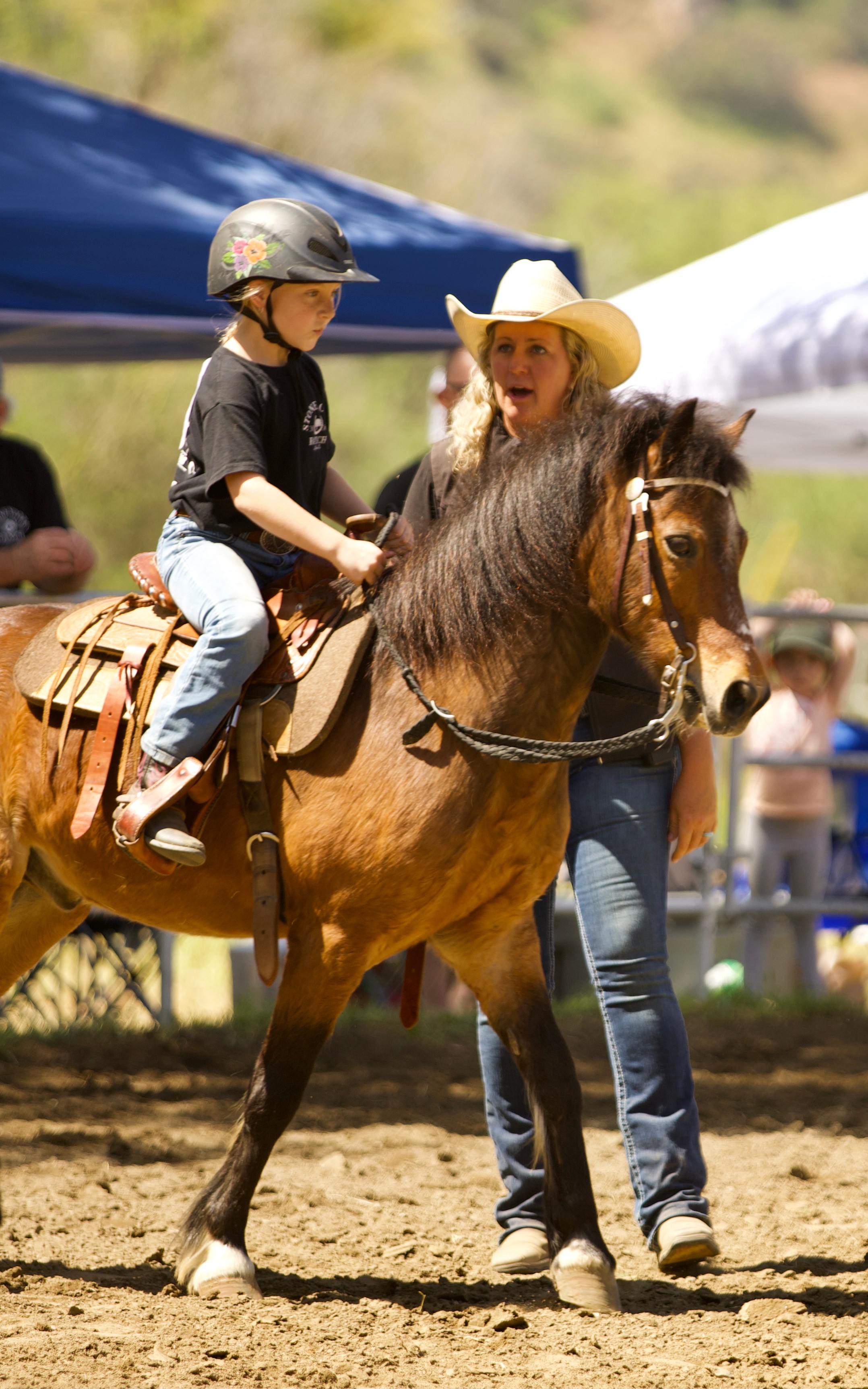 Child wearing a helmet rides a horse, guided by a woman in a cowboy hat, outdoors with tents in background.