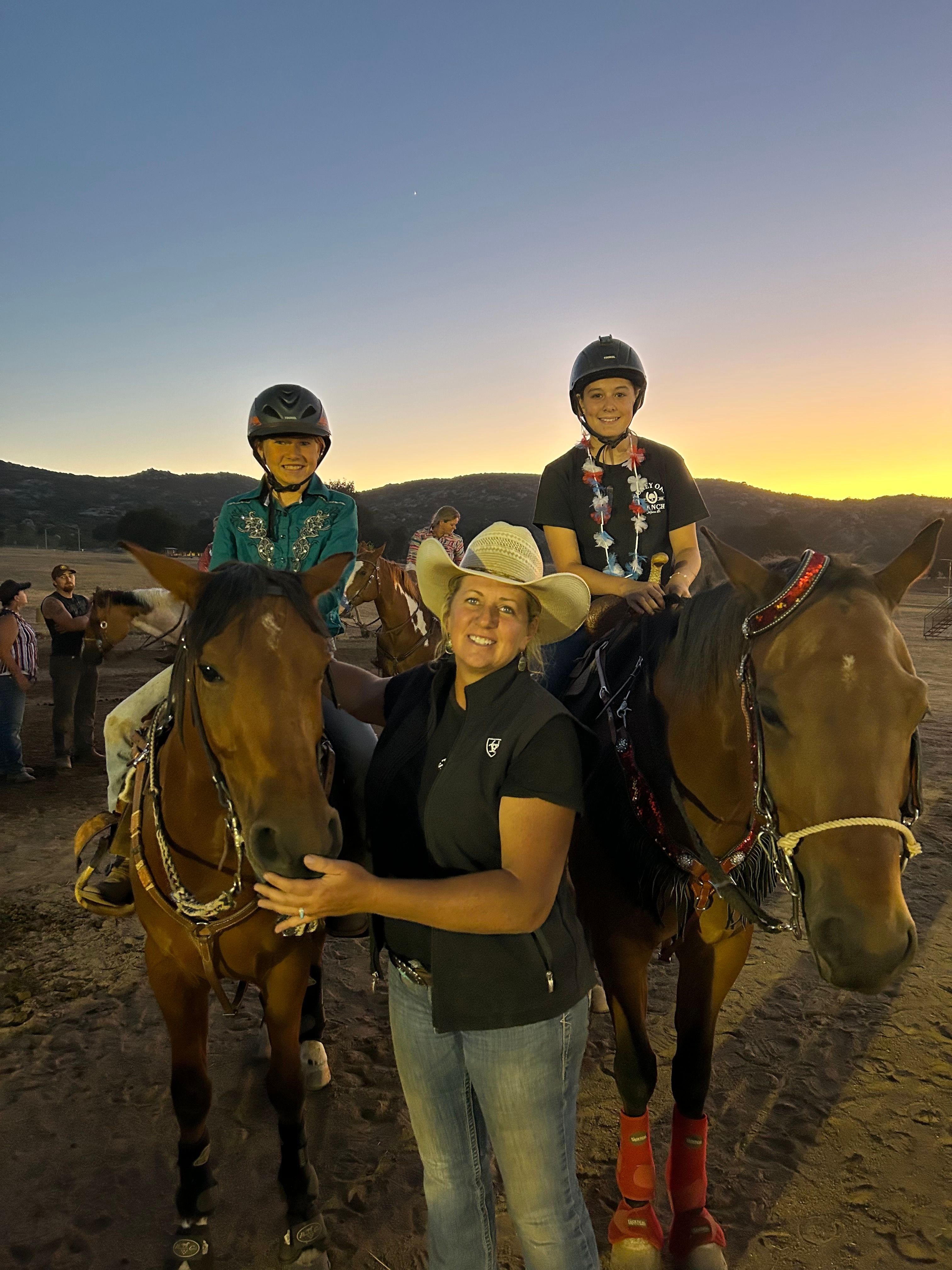 Four people standing outdoors during sunset, with a landscape background, some wearing jackets and hats, and one person petting a horse.