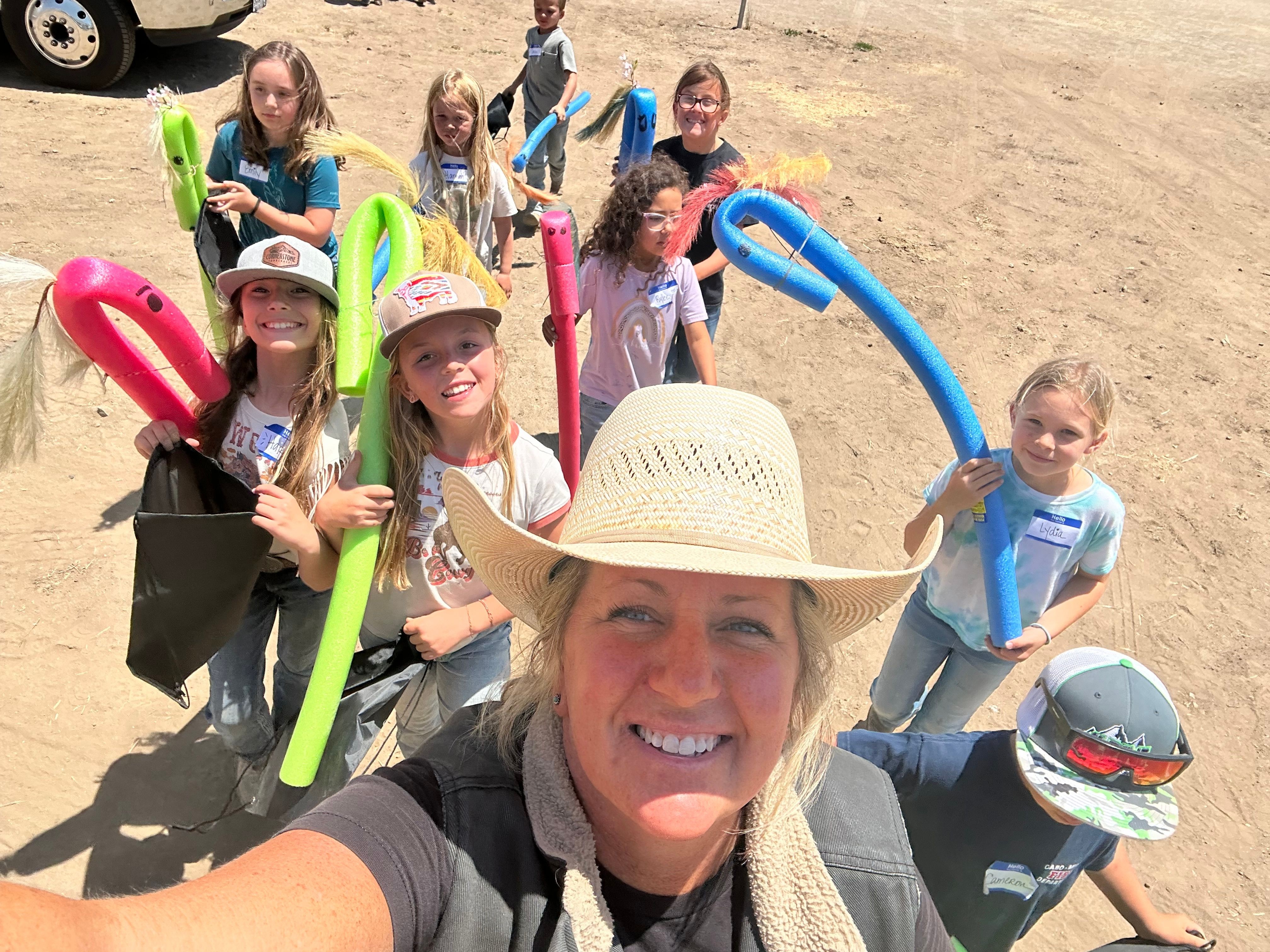Group of people on a sandy beach, smiling, holding colorful pool noodles and beach accessories, with a woman in the foreground wearing a hat.