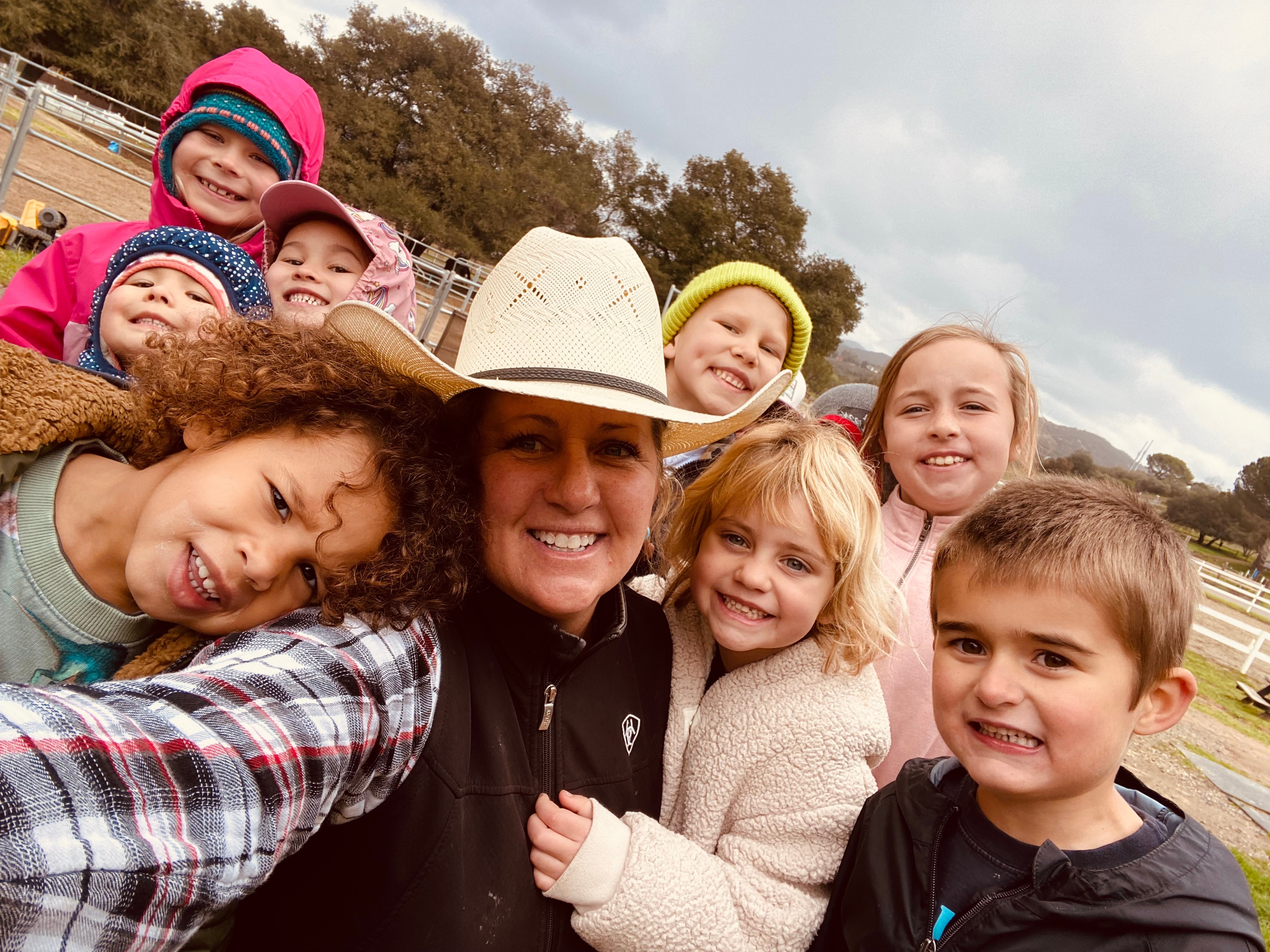 Group of children and an adult outdoors, smiling, wearing colorful hats and jackets, with trees and cloudy sky in background.